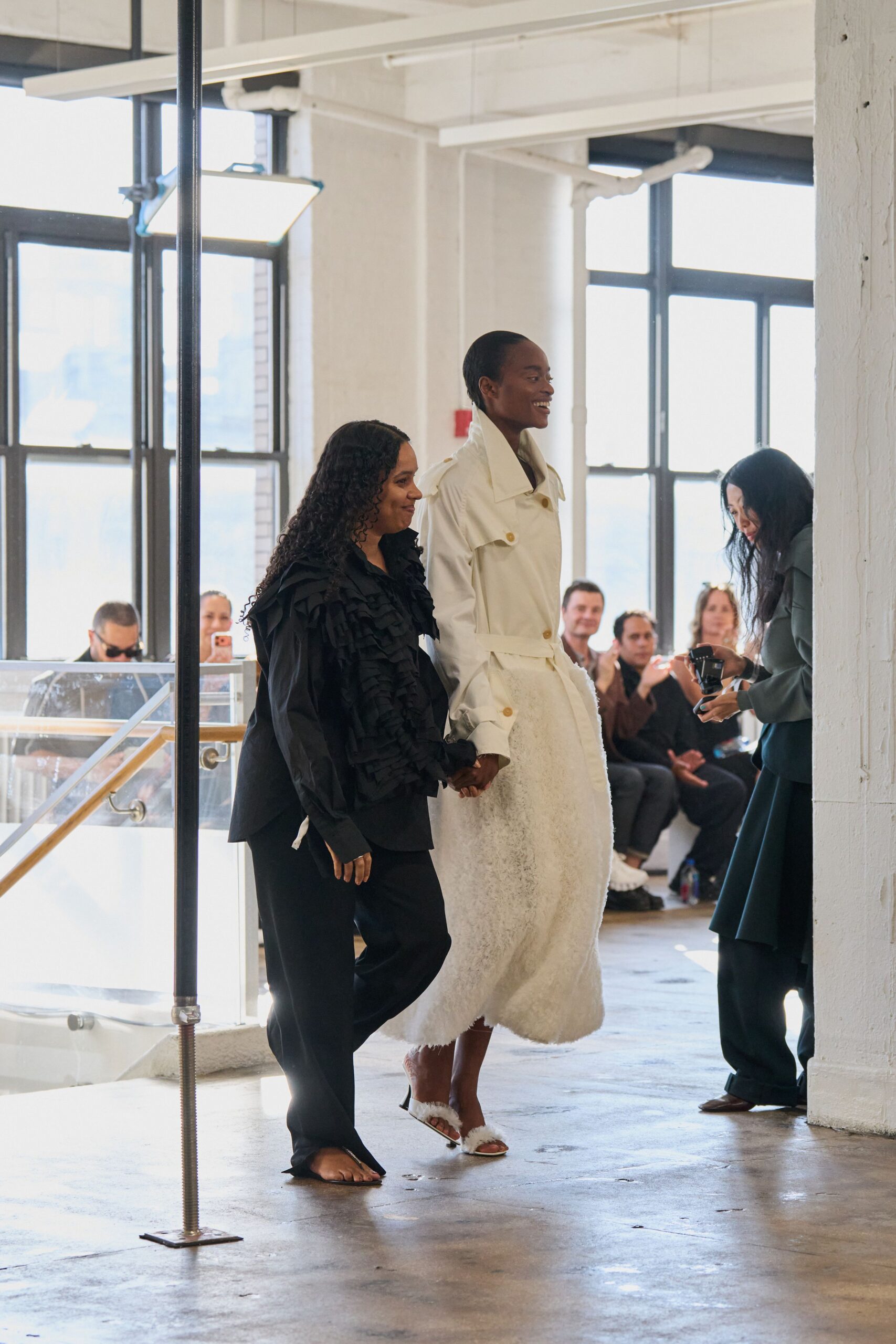 White trench coat with a high stand collar and horn-tone buttons. Long ivory skirt in fuzzy looped pile, hem brushing mid-calf. Open-toe mules trimmed with white fluff across the vamp. Applause in a sunstruck loft after somebody finally says thank you.