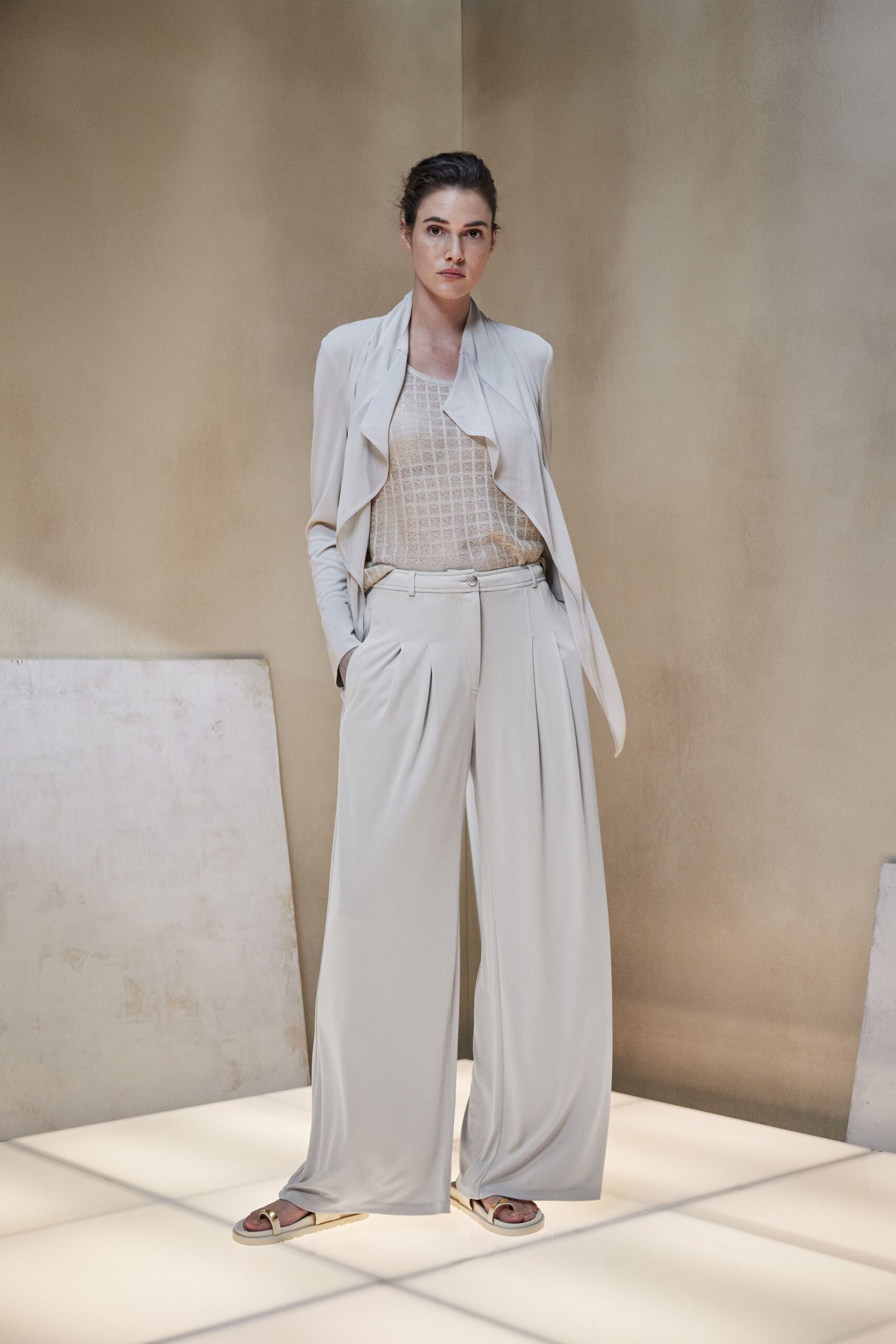 Pale stone jersey jacket with a soft waterfall front. Sheer ecru shell in open lattice knit, faintly shimmering. Fluid matching trousers with deep front pleats and a single button tab. Cream slides with a brushed gold toe strap. Walking across a tiled atrium before the building warms for the day.