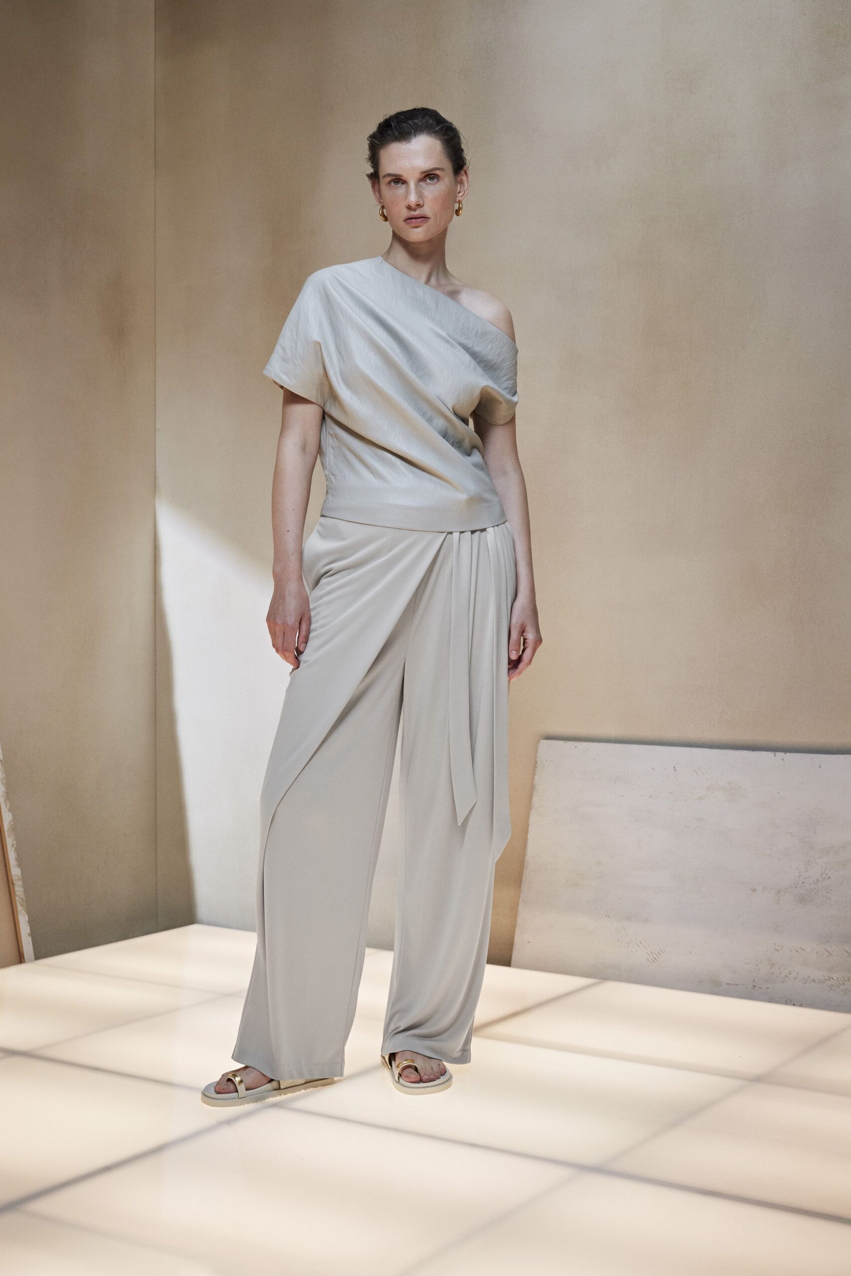 Pearl grey satin top draped off one shoulder, cuffed in a wide waistband. Matching fluid wide-leg trousers, gathered into a soft sash drop at center. Cream slides with brushed gold toe strap. Small gold hoops. Morning in a quiet hotel atrium before anyone claims the good chairs.