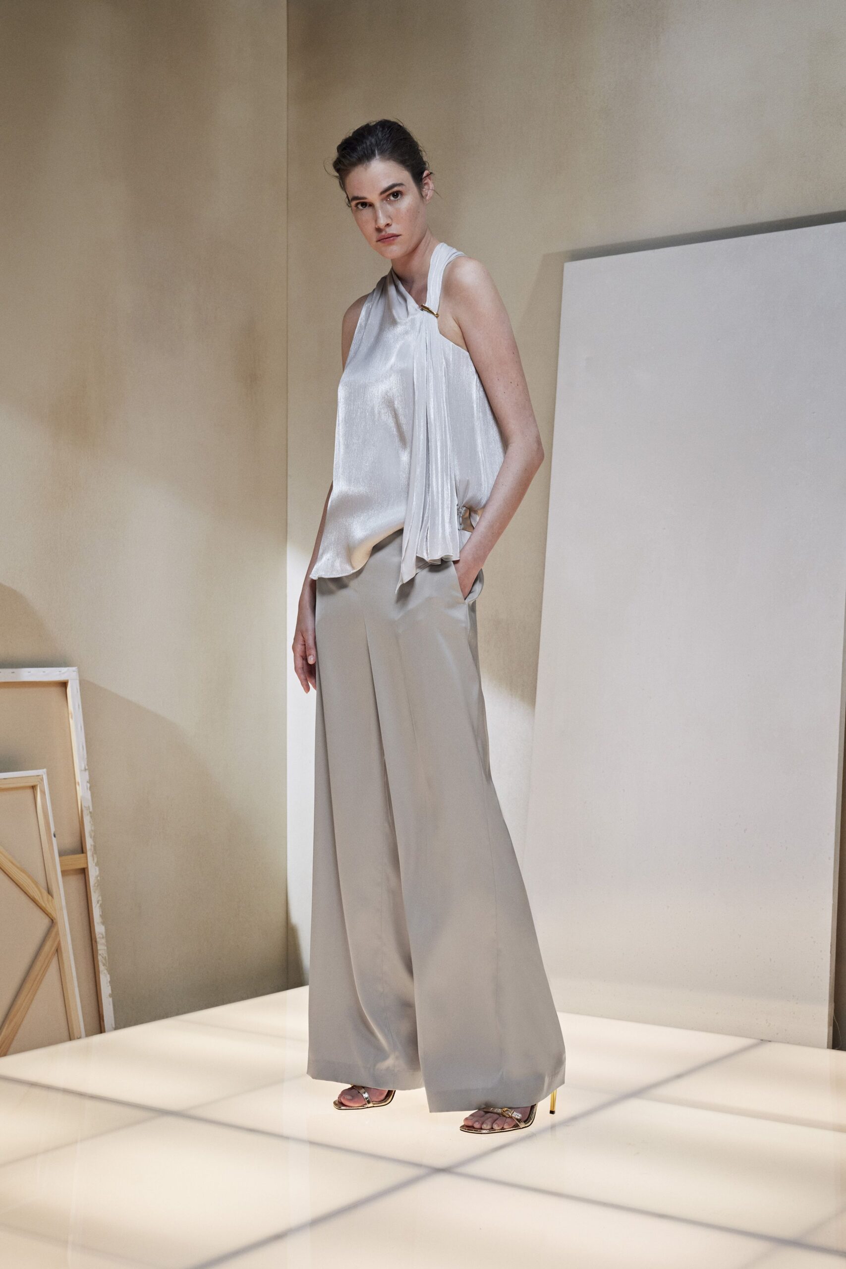 Pearly satin halter top with a draped panel pinned at one shoulder. Putty wide-leg trousers with a sharp crease and deep side pockets. Gold heeled sandals with slim metallic straps. At the apartment viewing where the floors are still covered in dust.