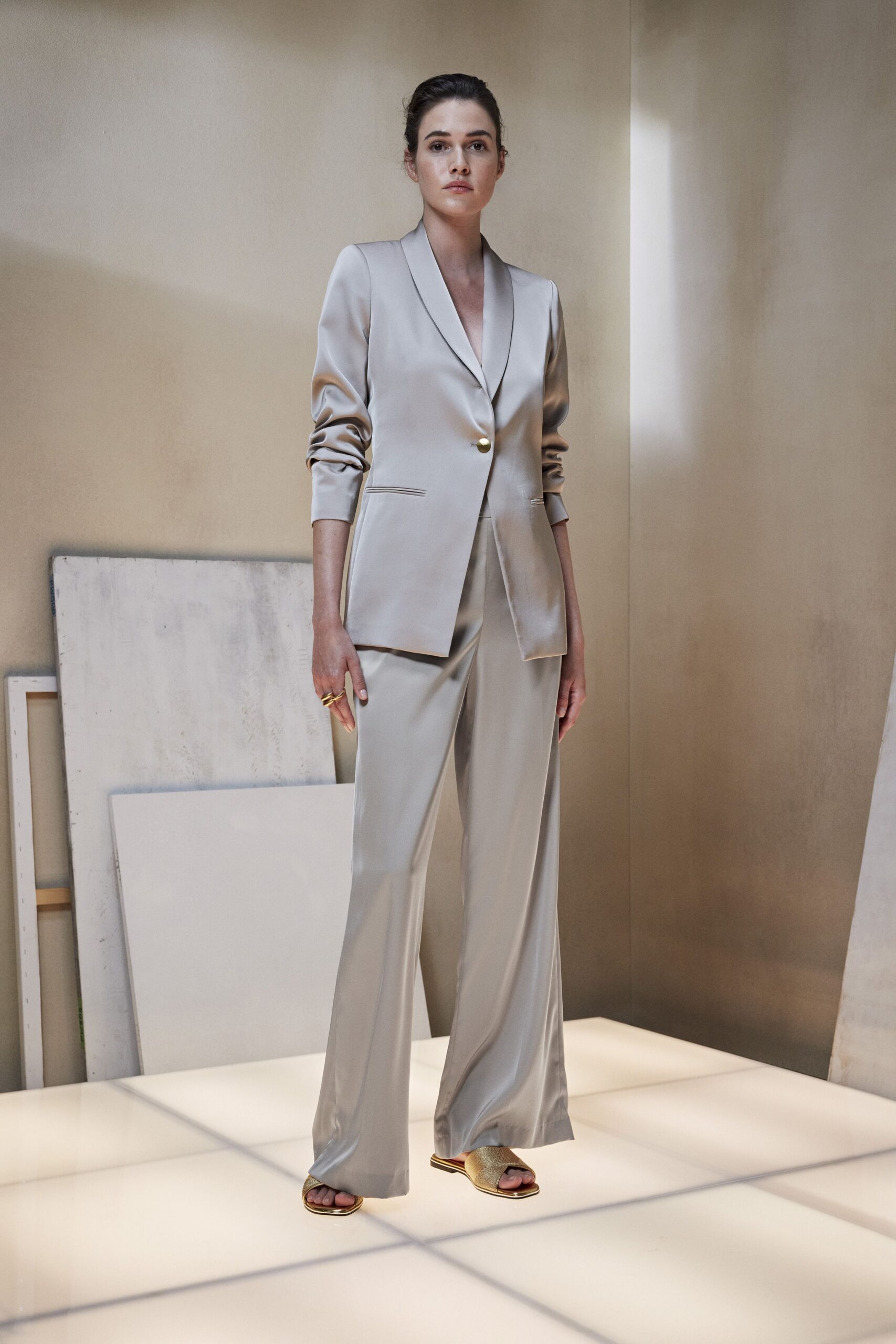 Pearl-grey satin blazer with a shawl collar and single gilt button. Matching fluid satin trousers, cut wide and breaking softly at the sandal. Gold slide sandals with a woven metallic vamp. Stacked gold ring looping across two fingers. For meeting the architect after hours in an apartment still full of drop cloths.