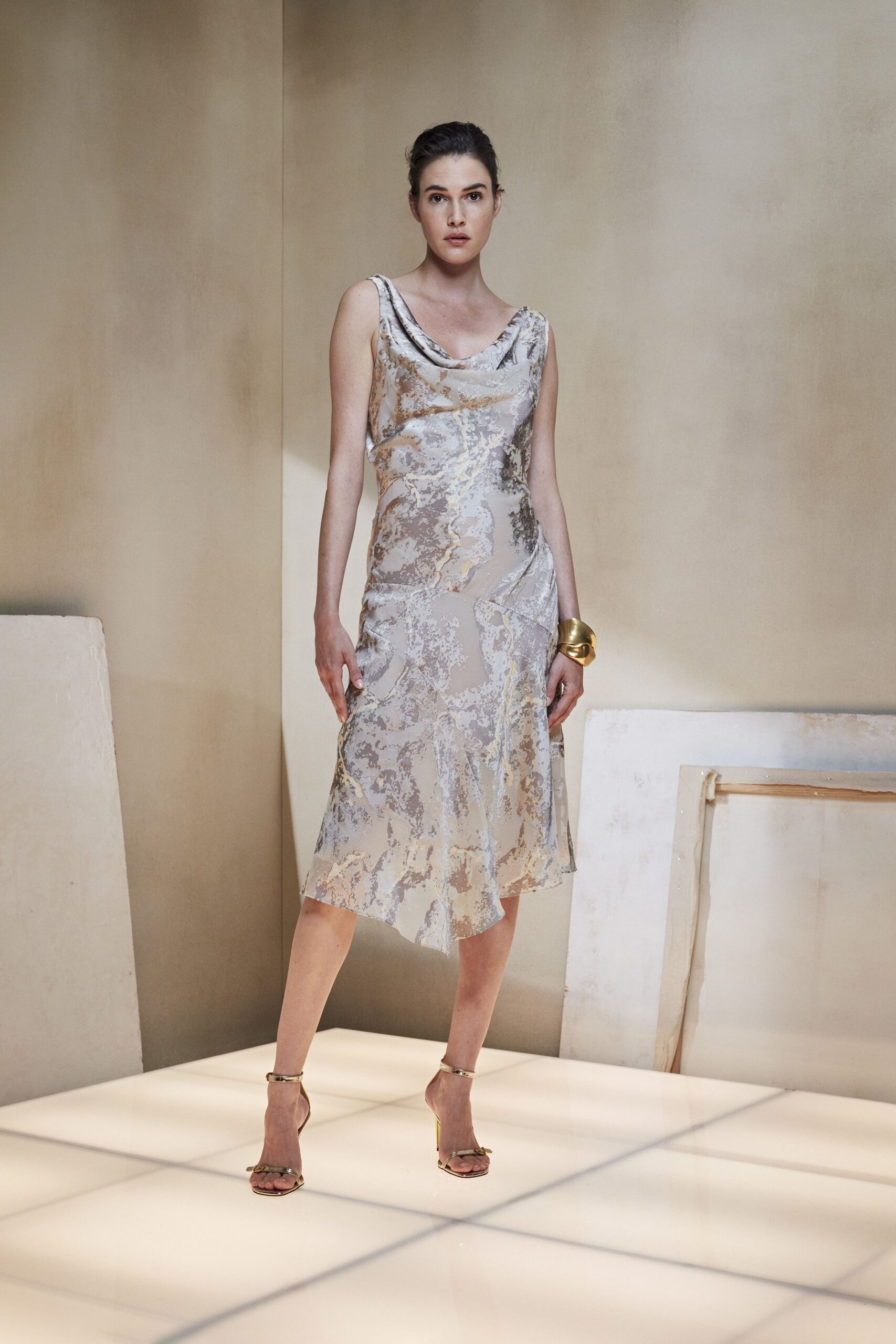 Bias-cut slip dress in cloudy silver jacquard, cowl neck and uneven hem. Wide gold cuff, softly crumpled like folded sheet metal. Gold ankle-strap sandals with a tiny knot across the toes. Walking through a hotel atrium after the florist has already packed up.