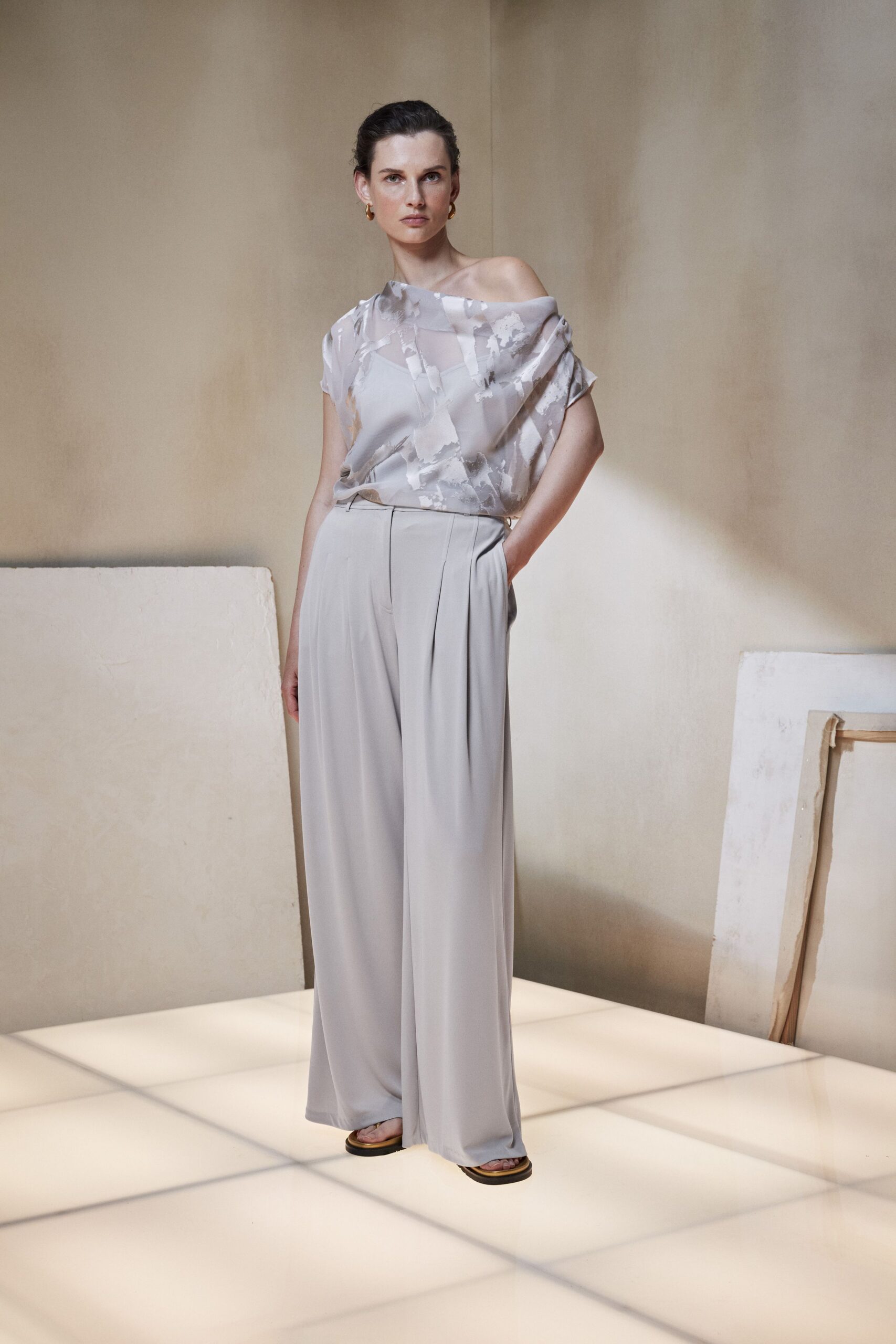 Misty grey satin top with a washed brushstroke print and one shoulder slipped bare. Pale dove wide-leg trousers, front pleats falling from a narrow waistband. Flat black-soled sandals. Small gold hoops. Early hotel breakfast, carrying your shoes back from the terrace.