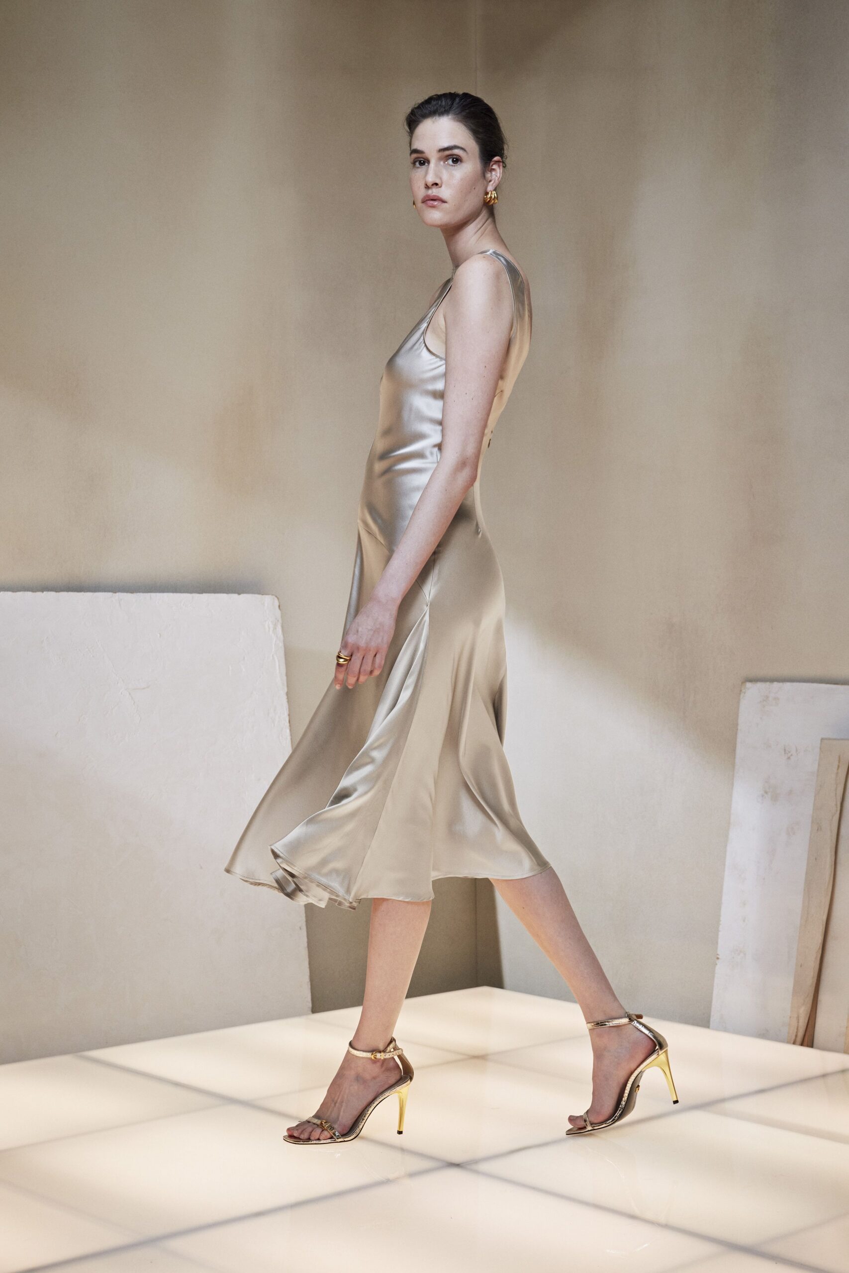 Champagne satin slip dress, bias-cut skirt kicking out at the calf. Small ridged gold hoops. Stacked gold bands. Gold ankle-strap sandals with mirror-bright stiletto heels. Crossing a hotel atrium before the wedding guests have come down.