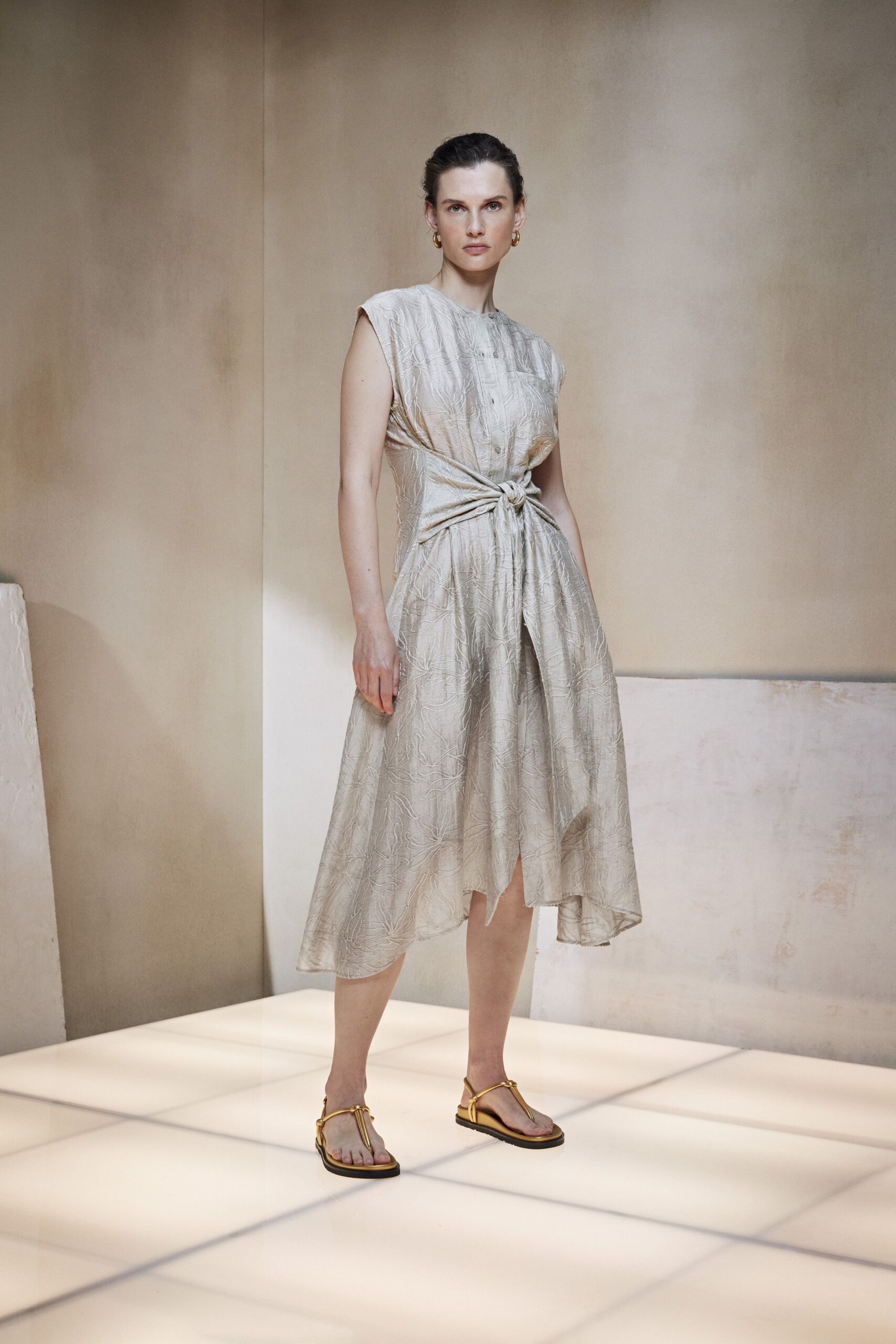 Small gold hoops at both ears. Pearl-grey jacquard dress with cap sleeves and a side-tied waist knot. Gold thong sandals with tubular straps over black soles. Garden ceremony on a heat-heavy afternoon, speeches not started yet.