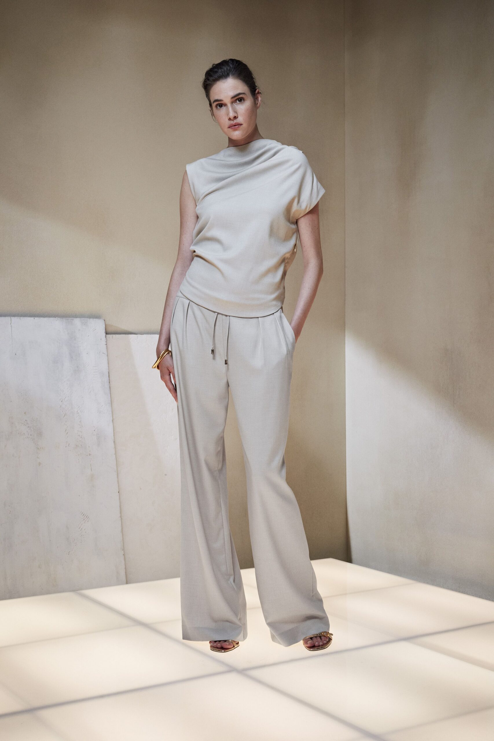 Ivory jersey top with a draped funnel neck and one cap sleeve. Pale stone wide-leg trousers, drawstring ends tipped in small metal aglets. Gold flat sandals with a knotted strap across the toes. Curved gold cuff. For crossing a hotel atrium before breakfast is being cleared.