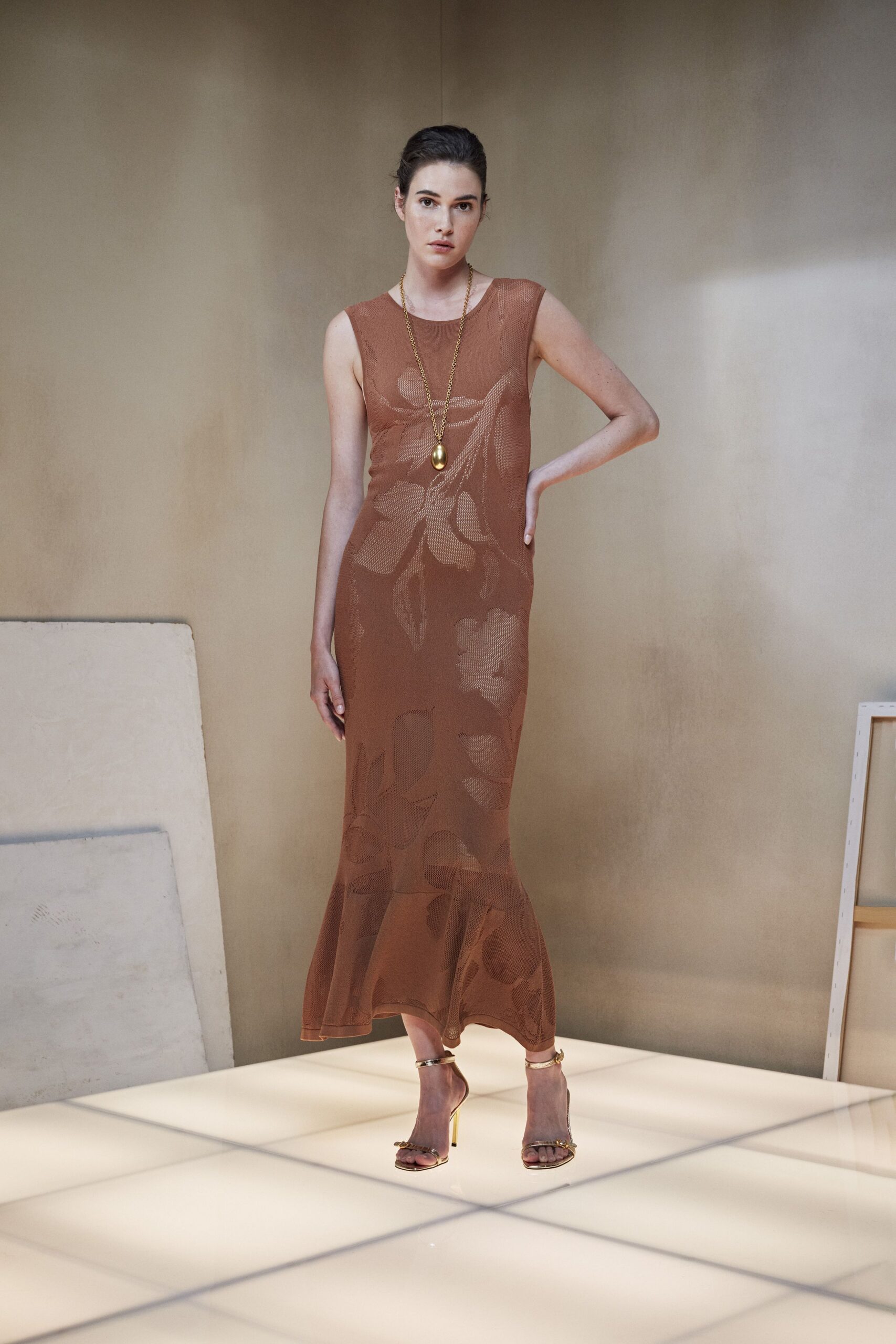 Long gold chain with a blunt egg-shaped pendant. Cocoa open-knit column dress, oversized floral pattern dissolving into the mesh. Gold ankle-strap stilettos with a tiny bar across the toes. At the hotel atrium after midnight, still warm from the terrace.