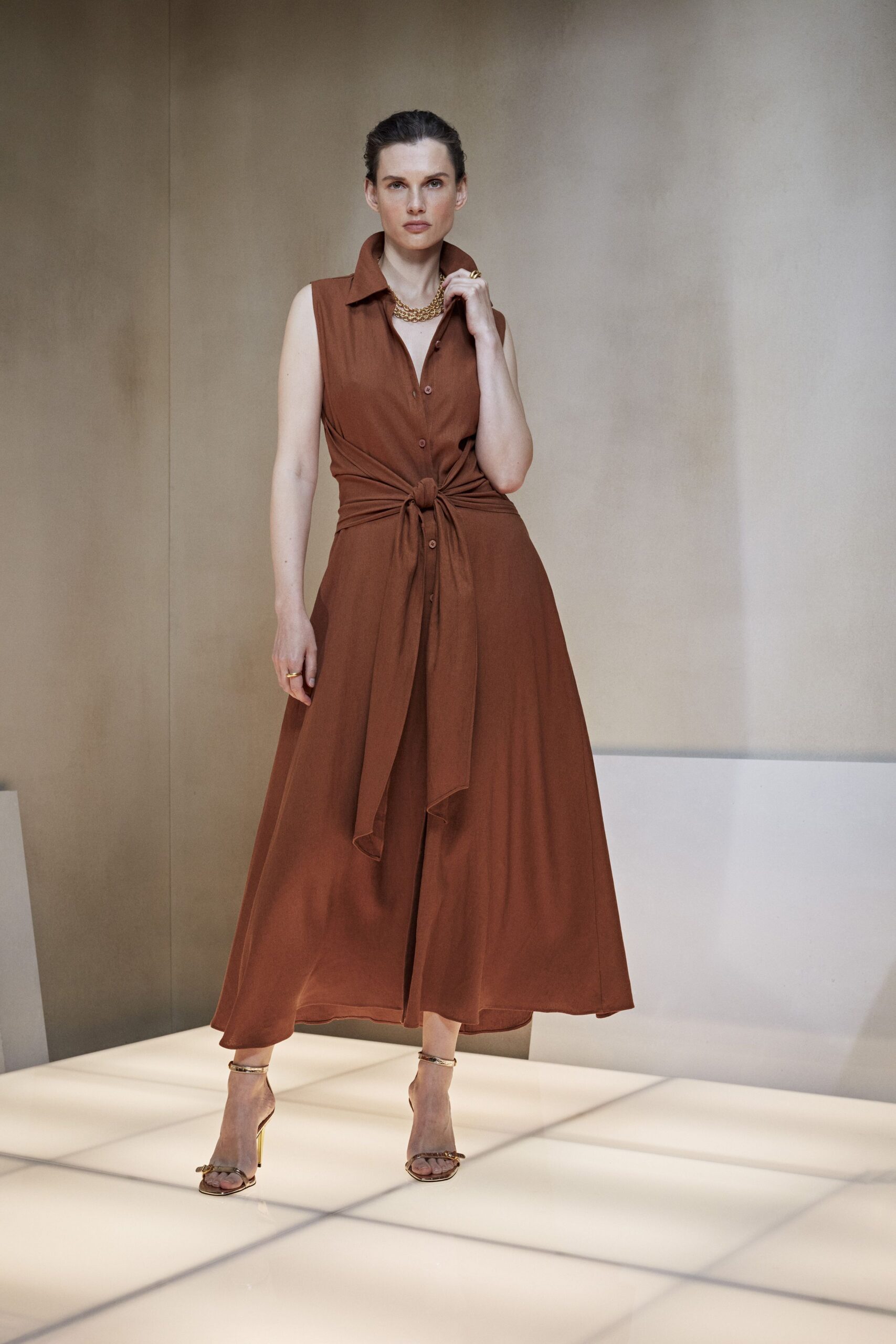 Russet sleeveless shirtdress in fluid crepe, waist tie knotted over the placket. Short double curb-chain in warm gold. Barely-there metallic sandals with slim ankle straps. Hotel atrium wedding, half an hour before the florist arrives.