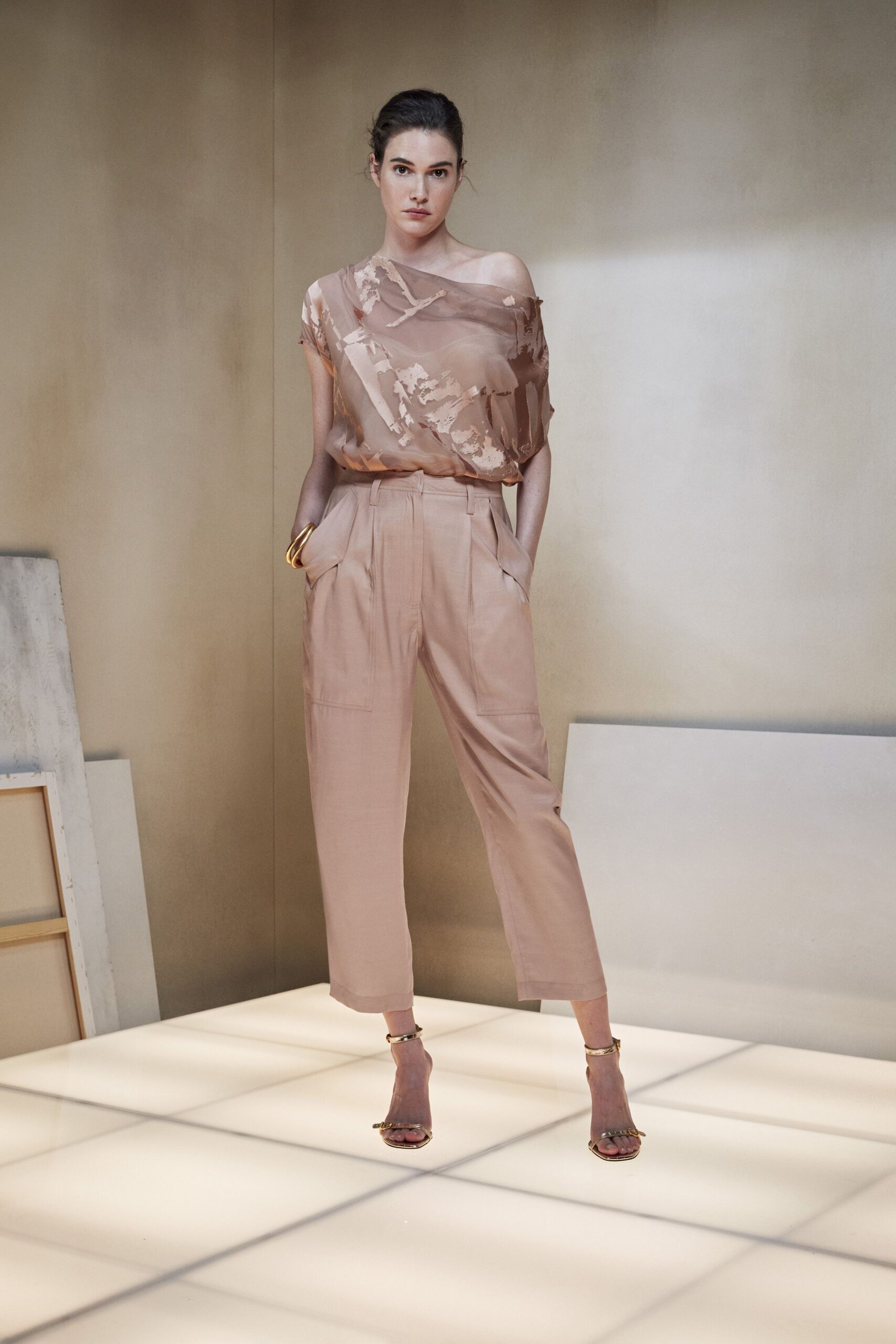 Sheer taupe chiffon top with a slouched one-shoulder neckline. Blush high-waisted trousers, front pleats and angled pocket flaps. Thick gold bangle at one wrist. Gold ankle-strap sandals with a tiny knot at the toe. Afternoon in a quiet apartment still smelling faintly of fresh paint.