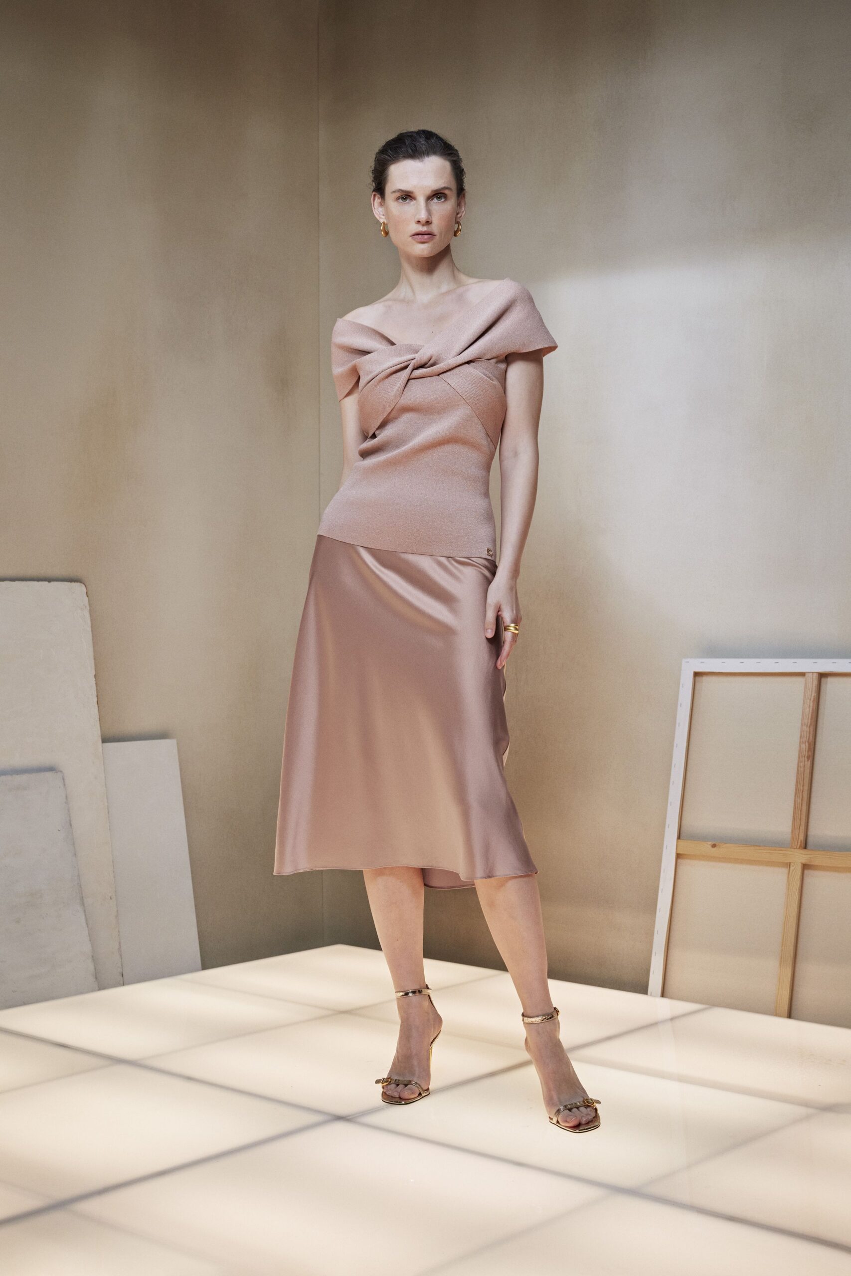 Blush knit top with a folded off-shoulder twist crossing the bust. Mink satin slip skirt, cut on the bias with a low glinting drape. Small gold hoops. Gold ankle-strap sandals, tiny bow glint at the toe. Sun still on the walls after a hotel wedding has ended.