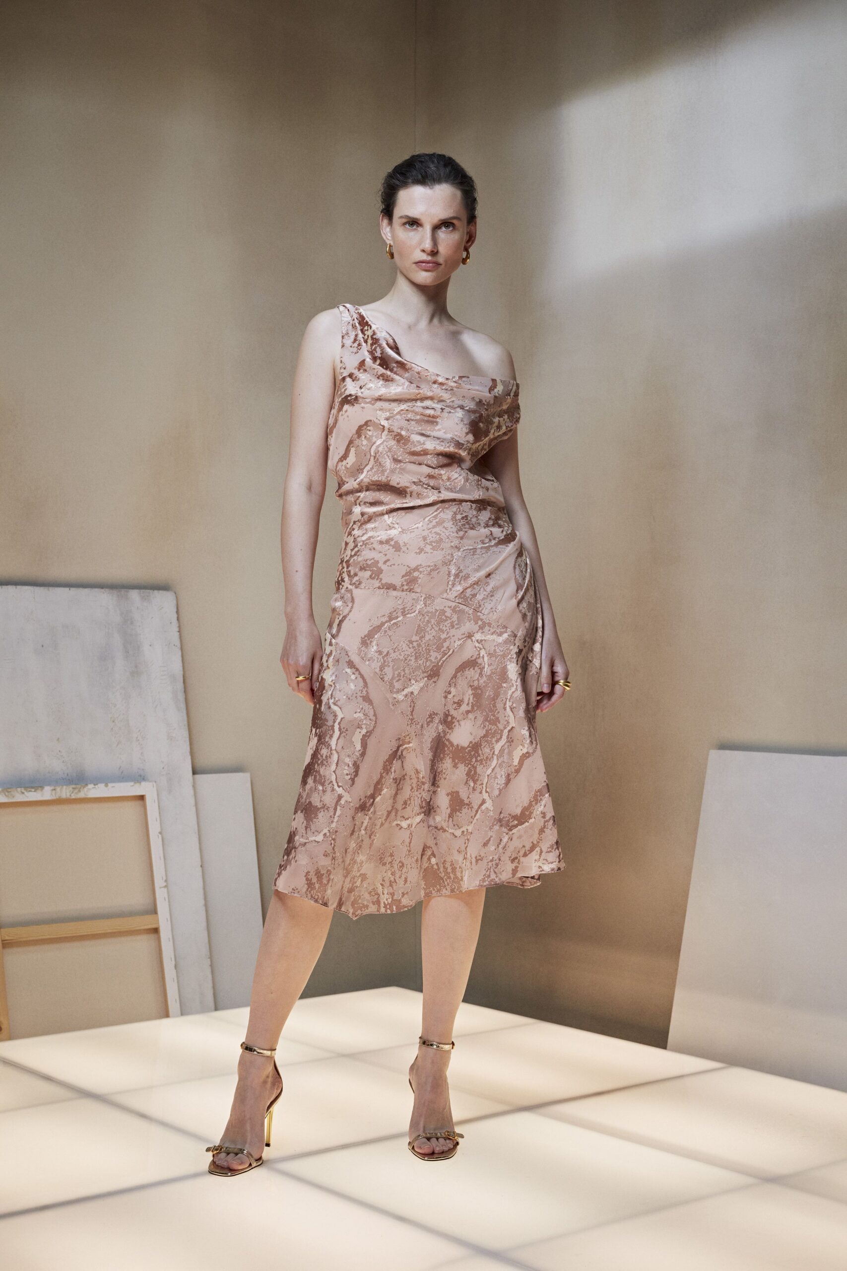 Blush satin slip dress with a draped one-shoulder neckline and marbled bronze print. Gold ankle-strap sandals with slim metallic heels. Small gold hoops. Mismatched gold bands. For a hotel atrium wedding that runs late into the champagne.