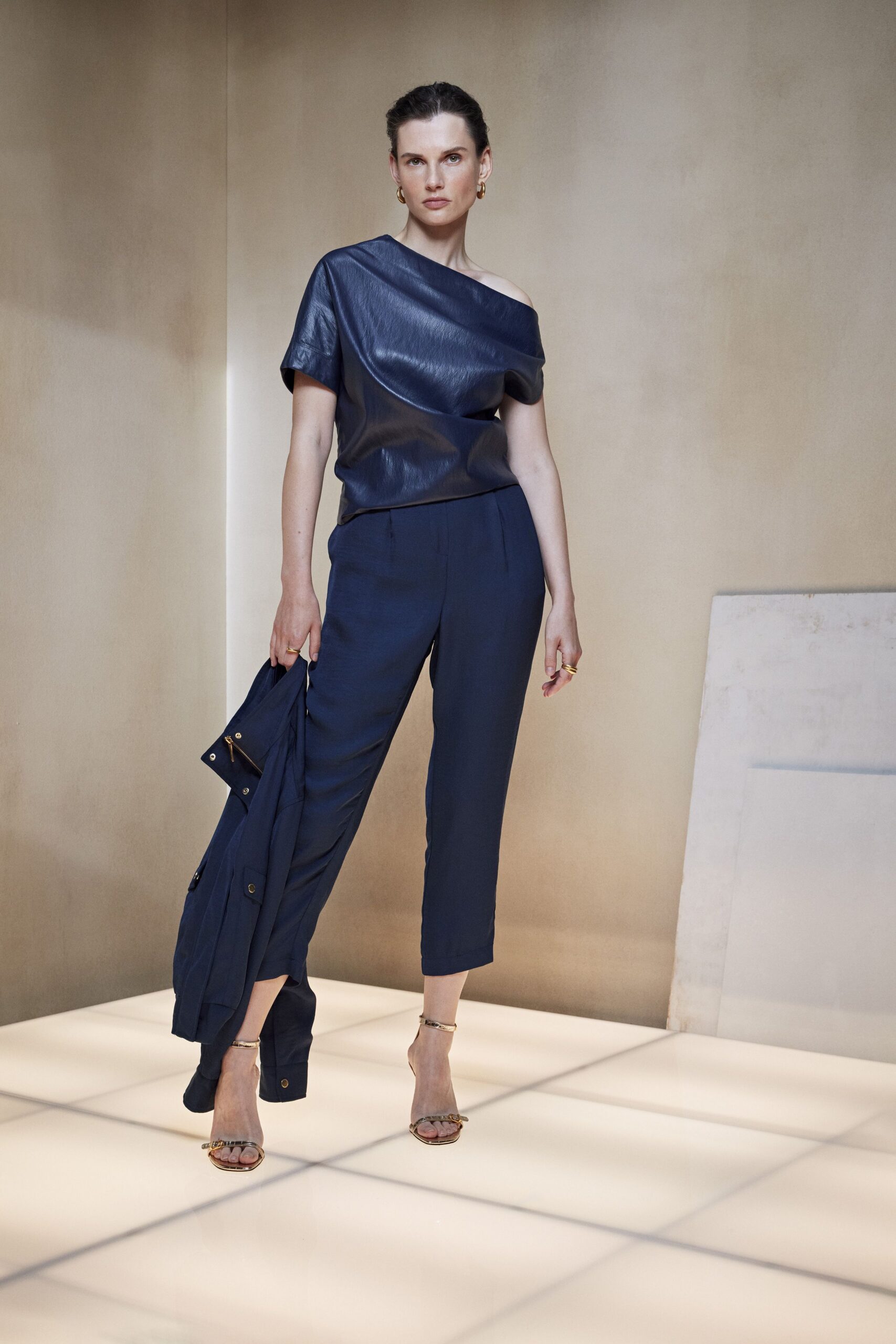 Ink-blue leather top draped off one shoulder in a folded crescent. Cropped navy trousers with a clean pressed front. Barely there gold sandals with chain across the toes. Matching coat carried low, brass snaps catching the light. For drinks on a hotel terrace before the air turns cold.