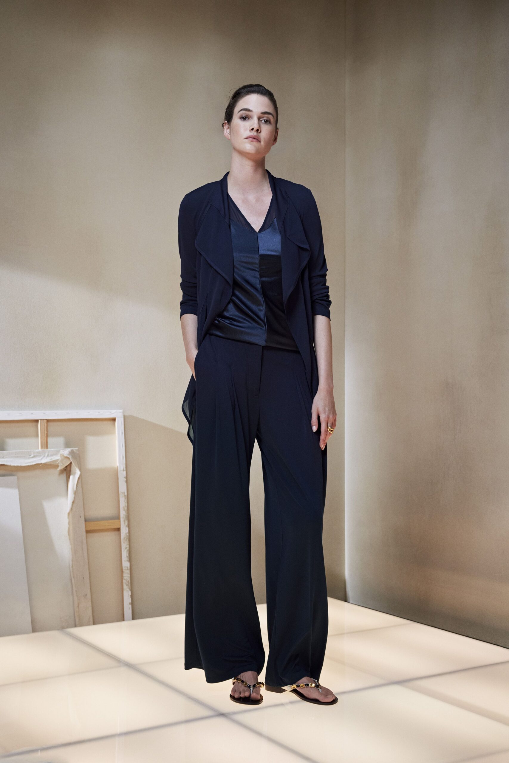 Ink jersey jacket with open waterfall fronts and pushed-up sleeves. Satin-faced V-neck top pieced in inky tonal panels. Fluid navy trousers pooling softly over the instep. Flat thong sandals with amber-studded straps. Chunky gold ring. For walking through a quiet hotel lobby before breakfast service.