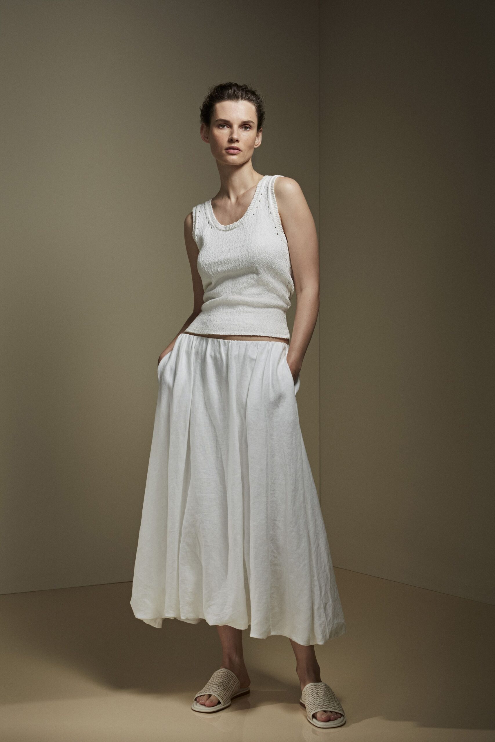 White boucle knit tank, tiny eyelets tracing the scoop and armholes. Full white skirt in airy washed linen, dropped from a low gathered waist. Cream woven slides with thick basket-plait straps. For crossing a tiled courtyard before the laundry has fully dried.