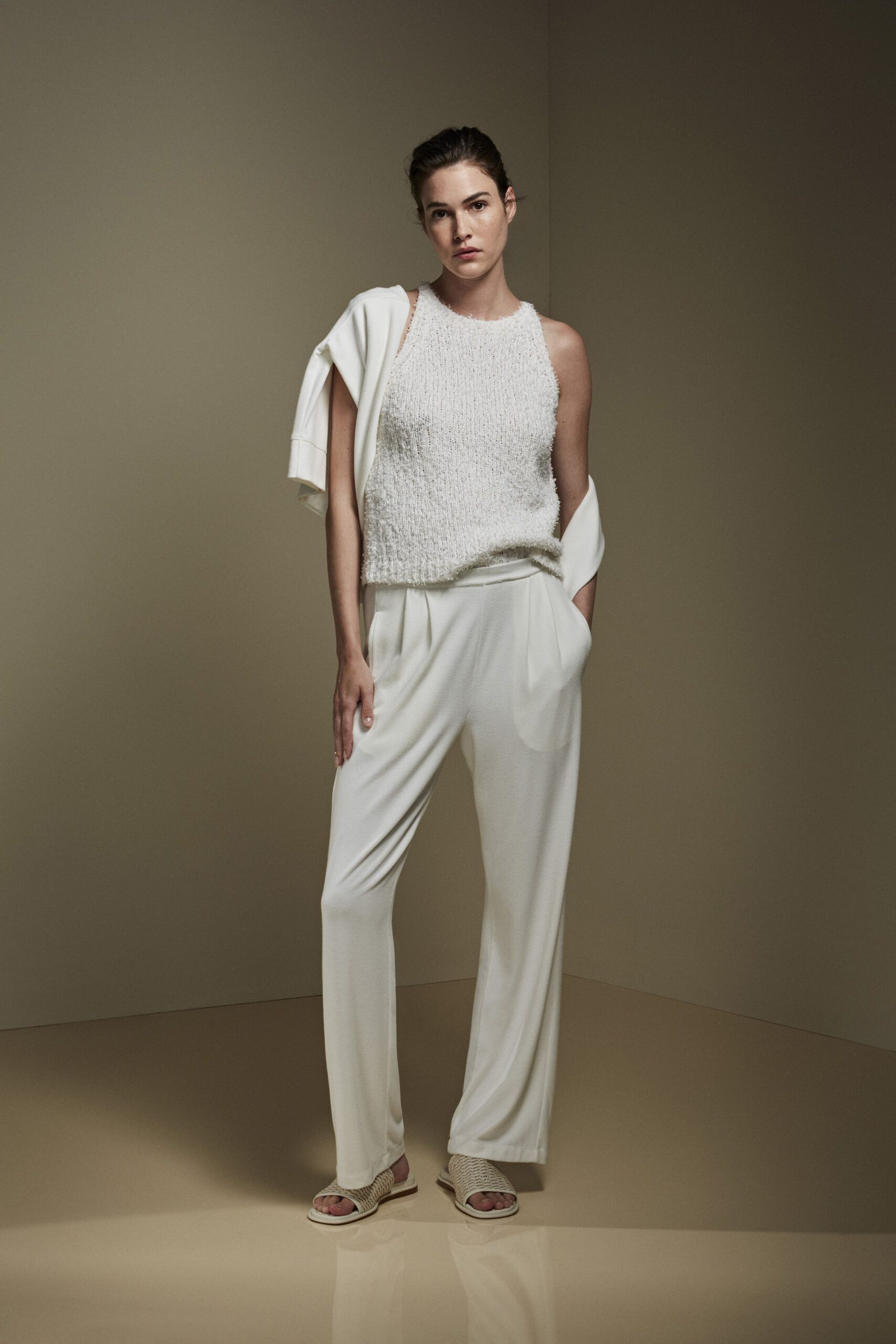 Ivory boucle knit shell with a fuzzy halo and ribbed hem. Cream sweatshirt shrugged off both shoulders, sleeves hanging loose. Fluid ivory trousers with soft pleats dropping from a wrapped waist. Braided raffia slides on thick white soles. Walking barefoot-adjacent through a rental house before the coffee finishes dripping.