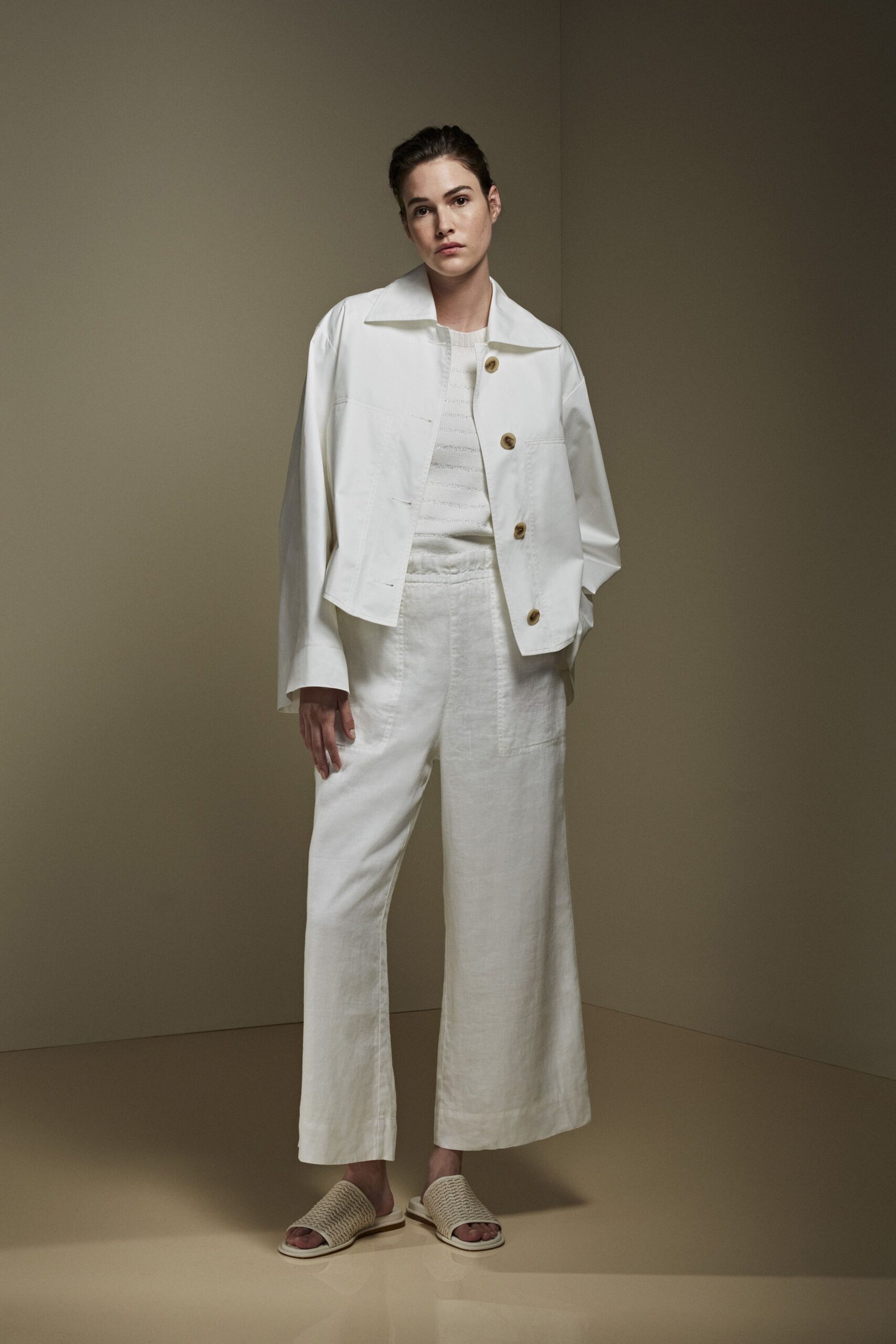 Cropped white jacket with a spread collar and oversized horn buttons. Ivory knit shell with faint horizontal laddered bands. White linen wide-legs with a soft elastic waist and cropped hem. Cream woven slides on thick flat soles. For the first coffee on a shaded hotel terrace, sandals still cool from indoors.