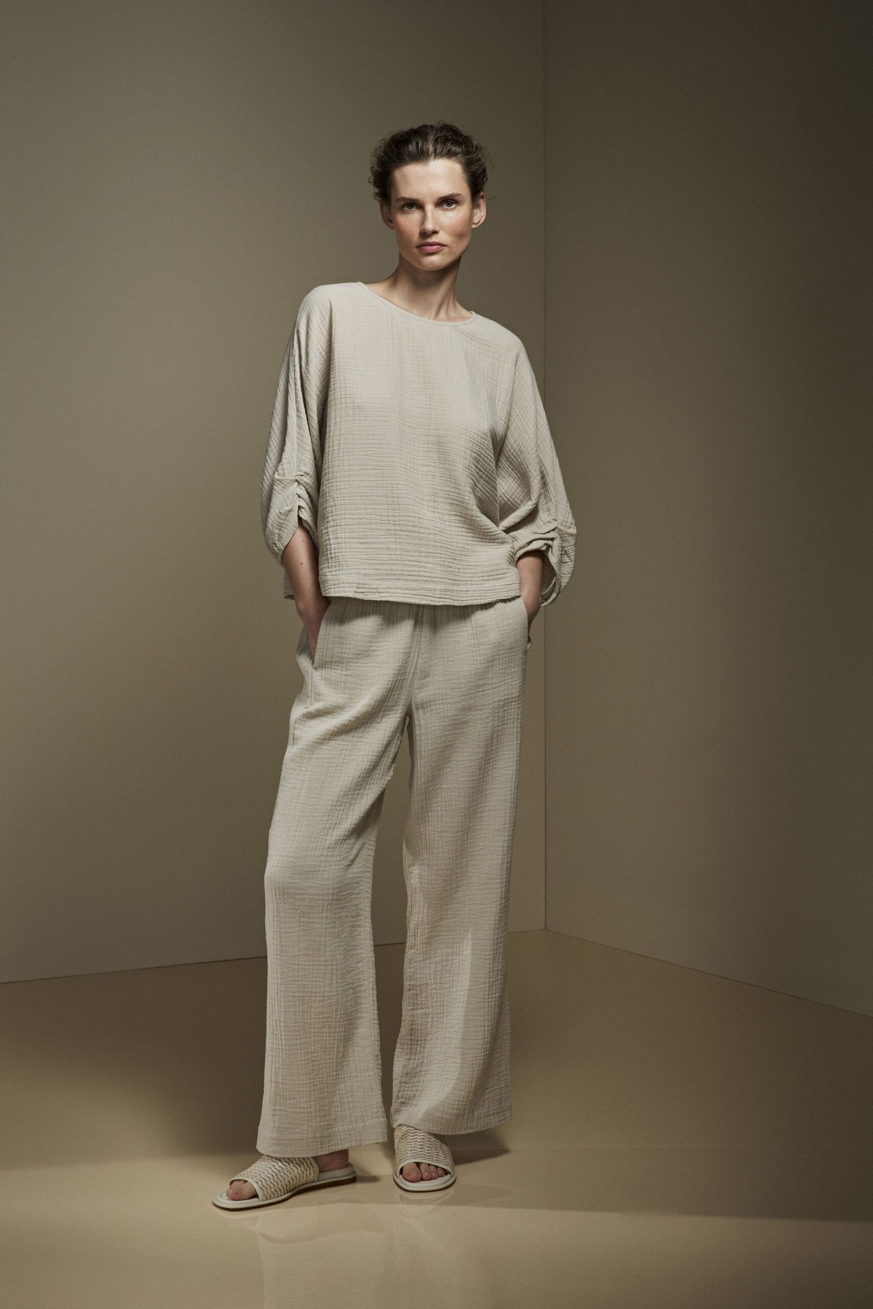 Oatmeal crinkled gauze top with ruched seams gathering the dropped sleeves. Matching gauze wide-leg trousers, soft pull-on waist hidden under the hem. Cream woven leather slides with a thick basket-plait strap. For the hour after a beach shower, windows open and the floor still cool.