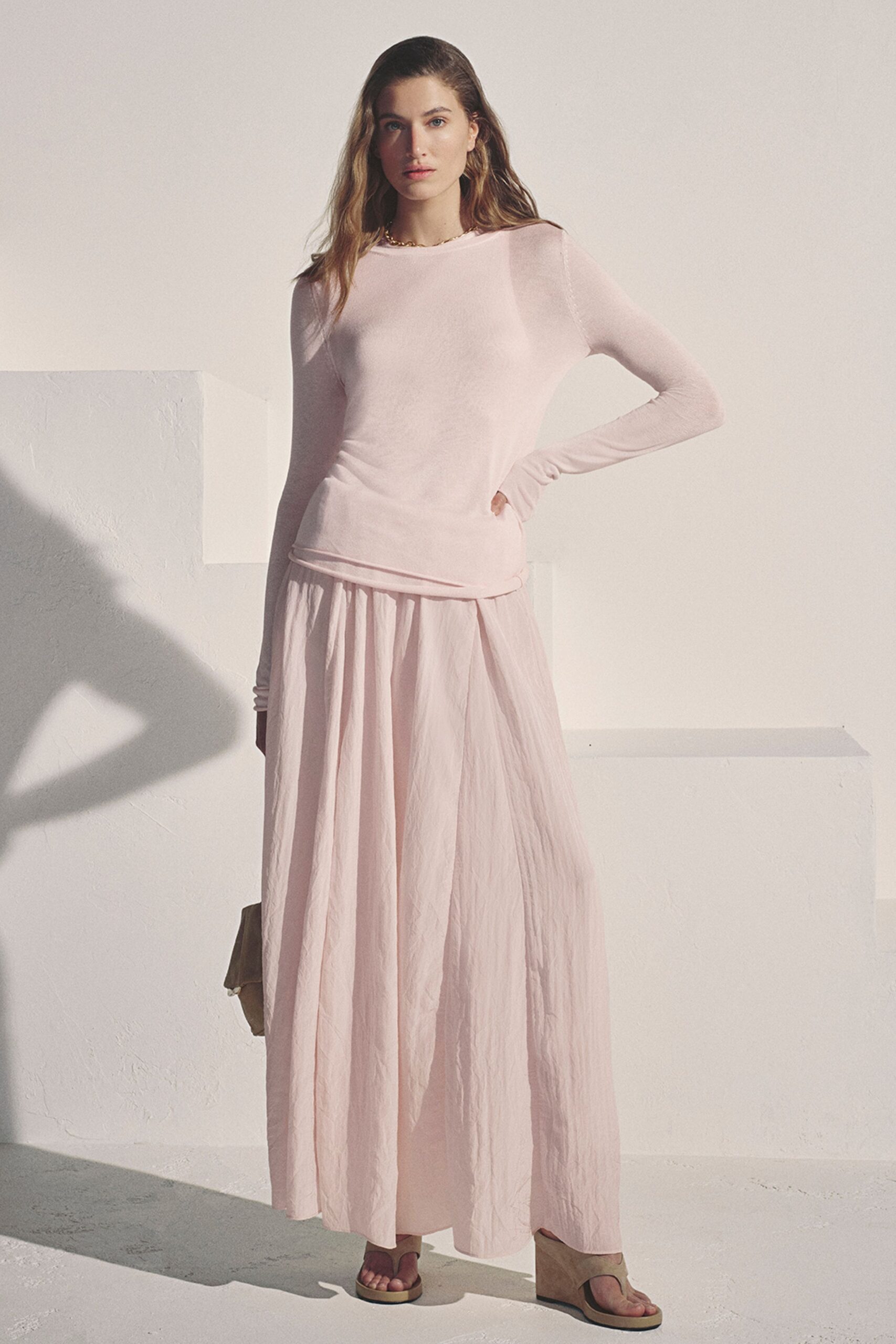 Short gold chain sitting close to the collarbone. Pale blush knit top with sheer shoulder seams and thumb-long sleeves. Crinkled blush maxi skirt, softly gathered into a dropped waist. Taupe suede thong wedges with thick platform soles. Sun through a stucco room, deciding whether to stay for one more coffee.