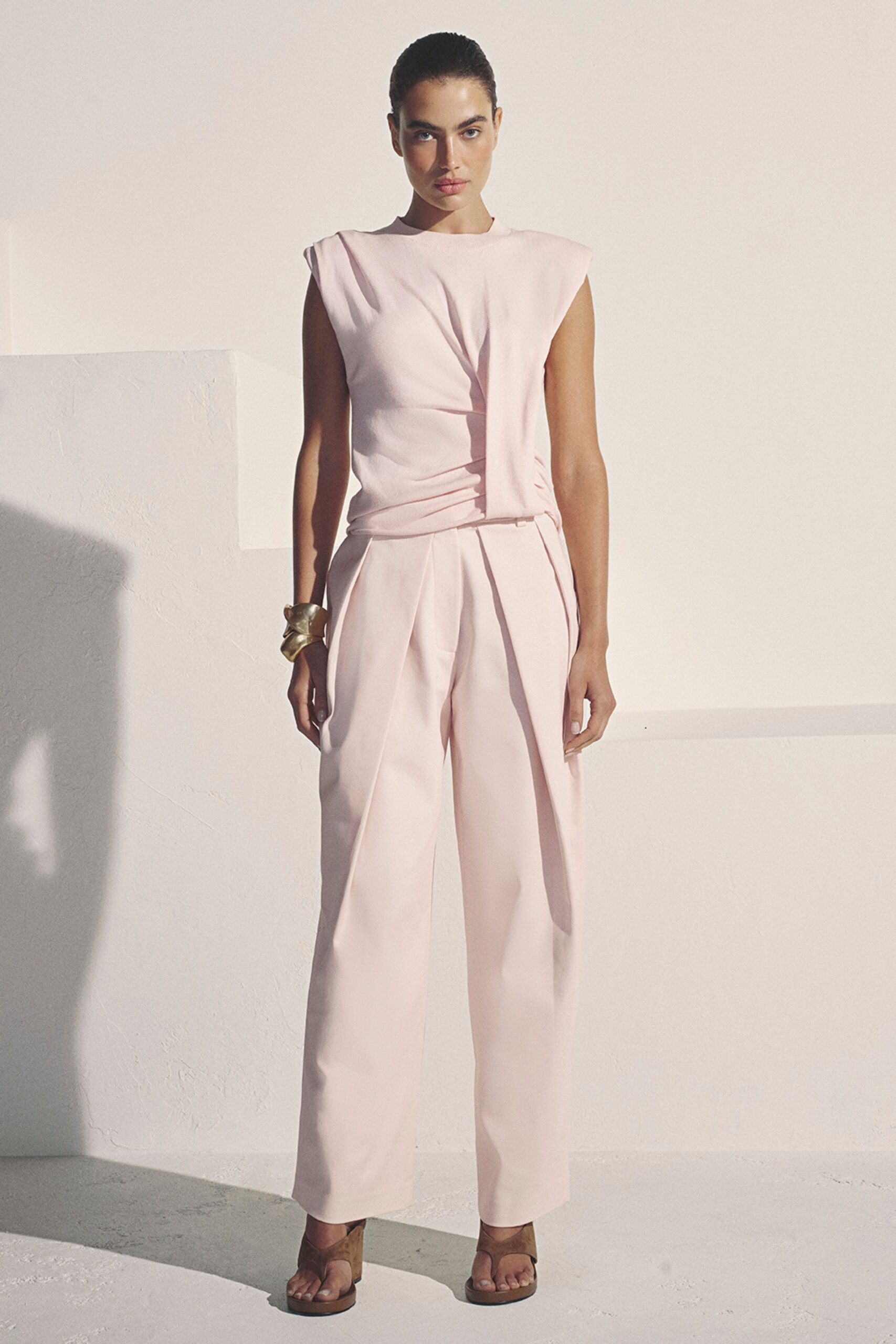 Blush crepe sleeveless top, twisted drape falling from the right shoulder. Blush crepe wide-leg trousers with a sharp front pleat. Crushed gold cuff, folded like soft metal ribbon. Tan suede thong sandals with thick block heels. Late afternoon in a whitewashed villa, shutters thrown open to the heat.