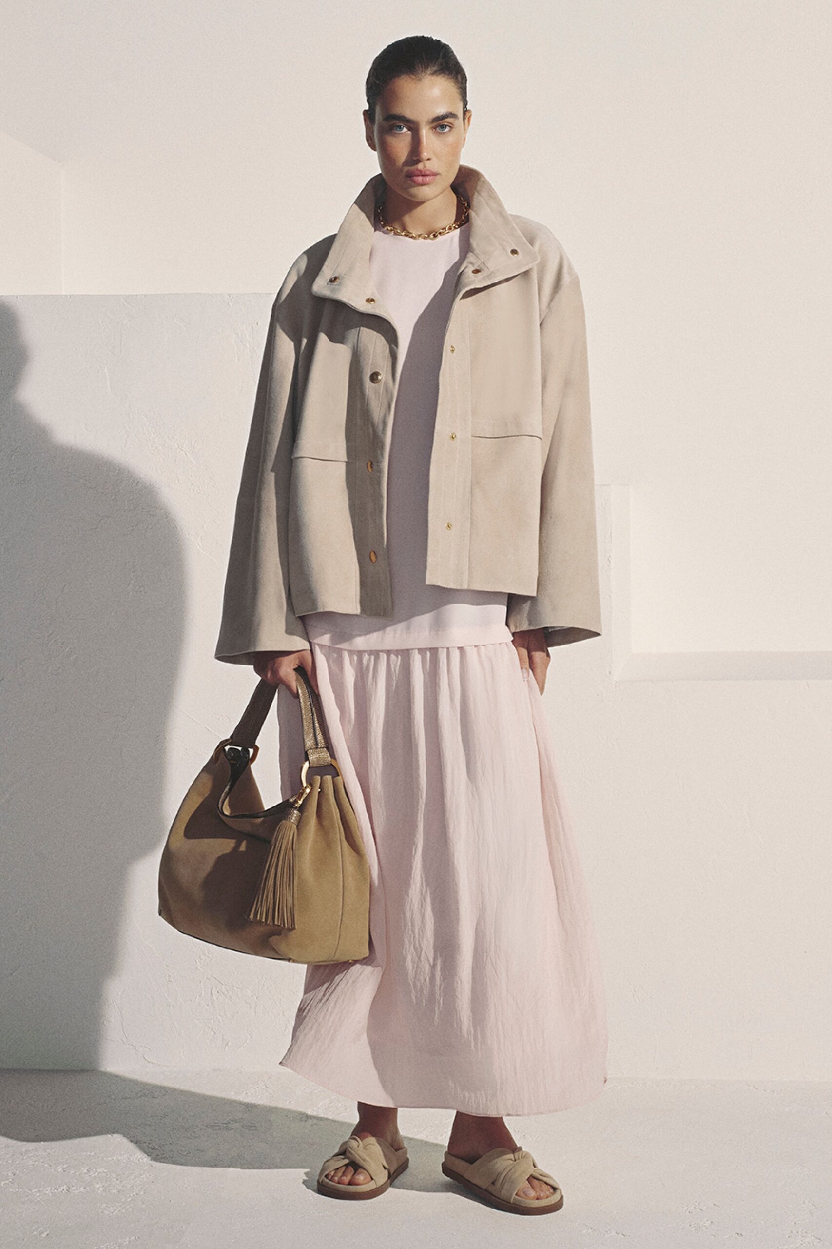 Pale stone suede jacket with a funnel collar and gold snaps. Blush crinkled dress, dropped waist gathered into a floaty skirt. Chunky gold chain at the collarbone. Taupe suede bucket bag with a long tassel swinging low. Cream leather slides with broad twisted straps. For picking up figs and good bread before the heat sets in.