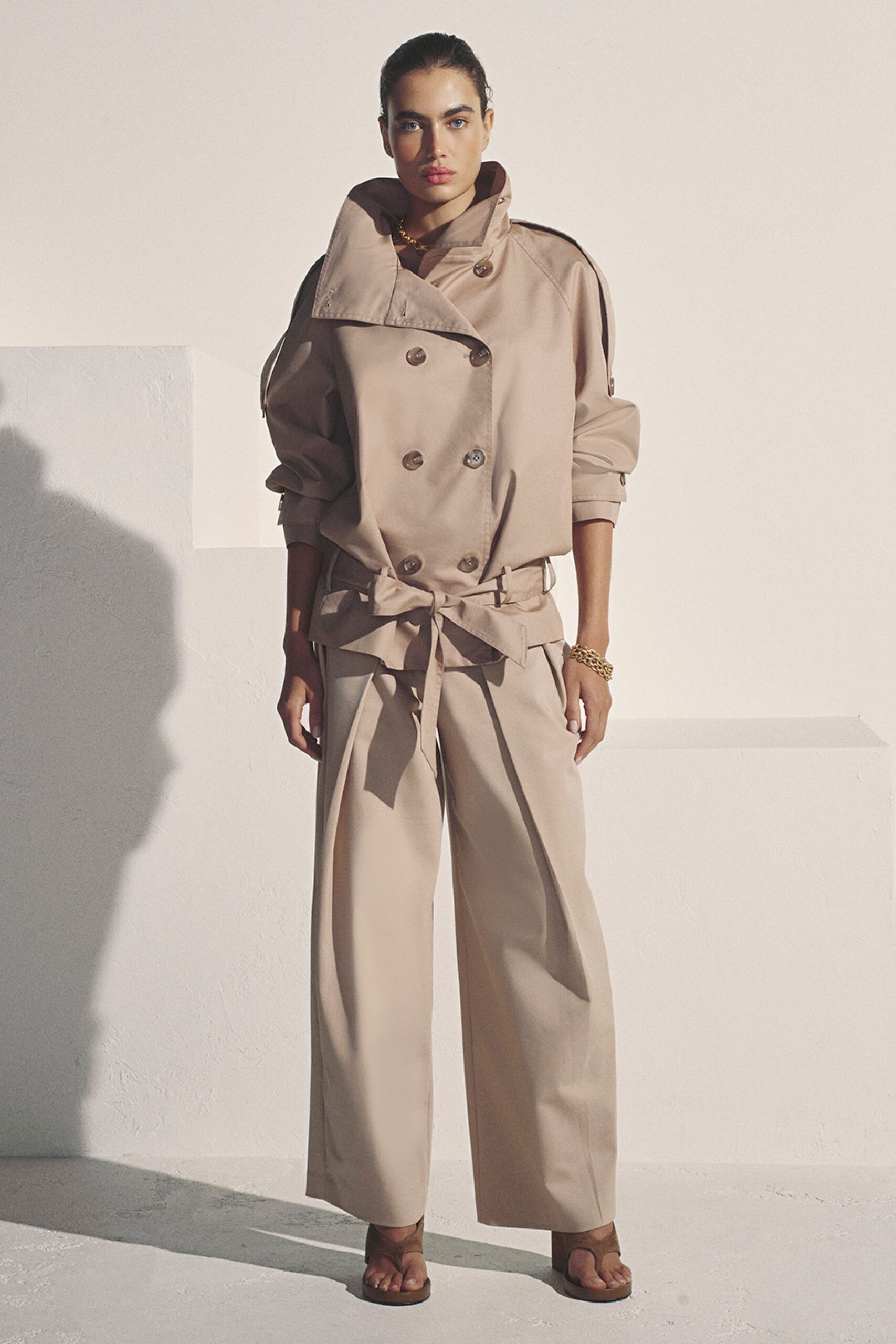 Sand cotton trench jacket, storm flap kicked high and sleeves strapped back. Chunky gold curb links at the throat. Matching wide-leg trousers with a long self-tie belt hanging past the fly. Twisted gold chain bracelet. Brown leather thong sandals with a square sole. For crossing a hotel terrace before the heat settles in.