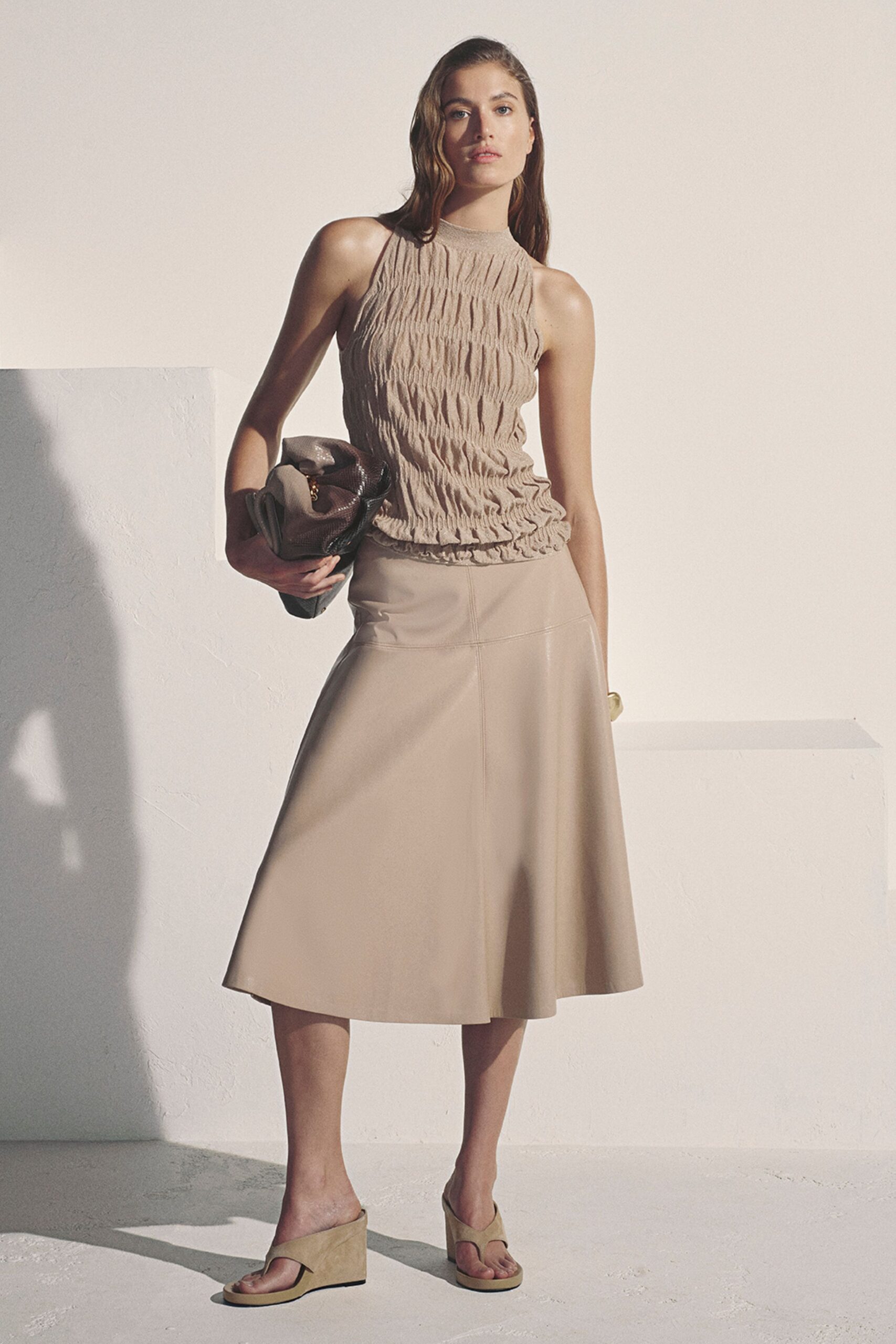 Sand jersey halter top densely ruched into vertical channels. Folded taupe snakeskin clutch with a small gold ring peeking out. Putty leather midi skirt, paneled seams opening into a soft flare. Beige suede thong wedges. For the hotel terrace just before the heat finally breaks.