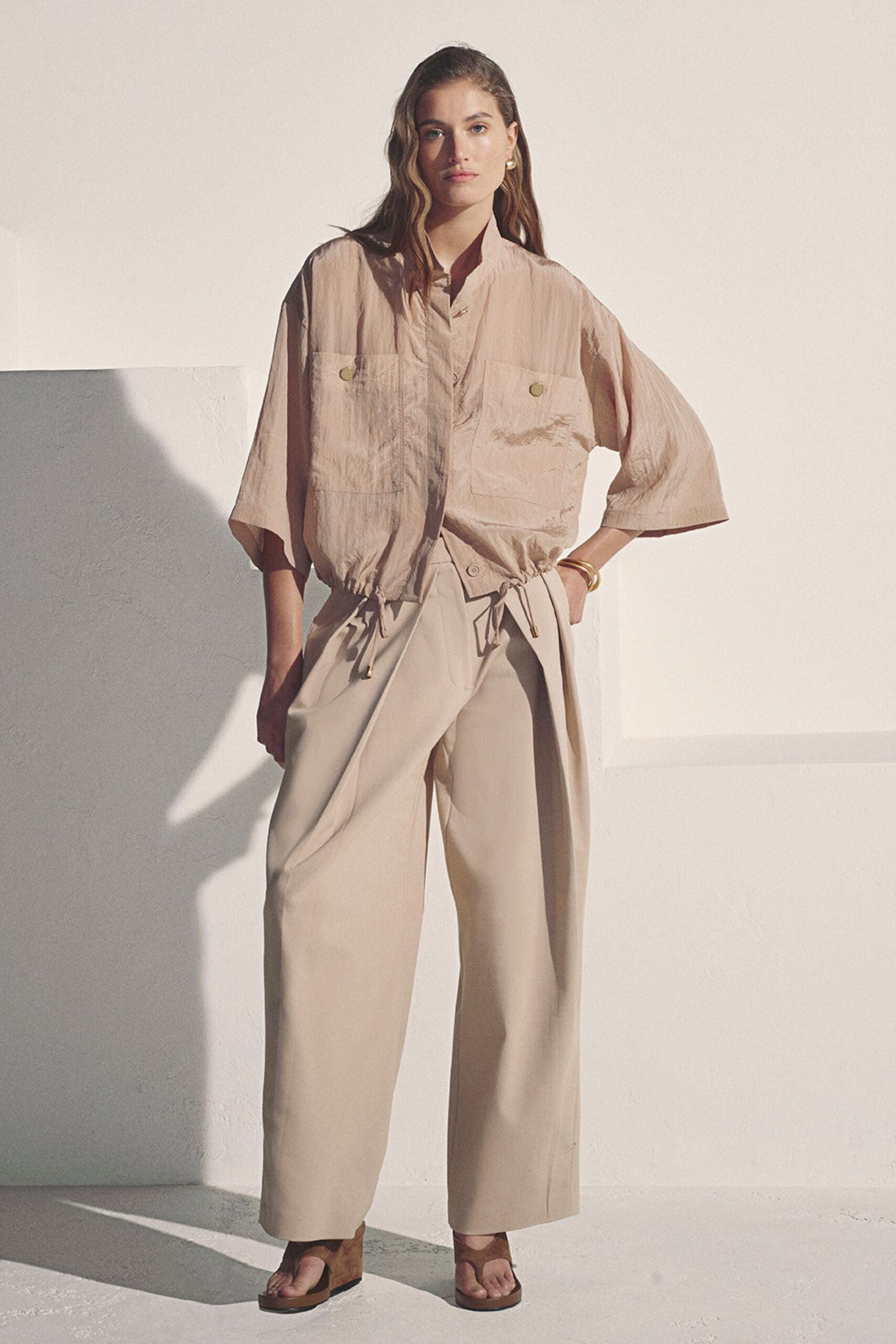 Small gold hoops catching the side light. Blush crinkled fabric blouson with oversized patch pockets and drawcord hem. Stone wide-leg trousers with deep pleats and a wrap-front overlap. Stacked gold bangles. Brown suede platform thong sandals. For the hour before dinner when the terrace still holds the day's heat.