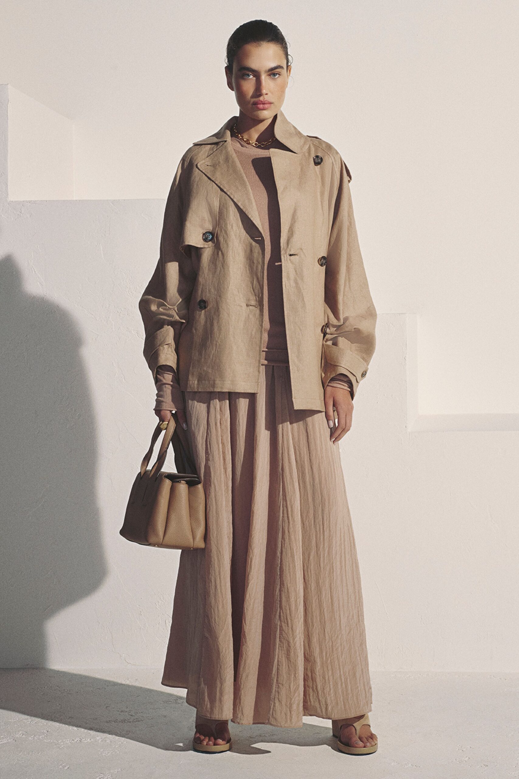 Sandwashed beige trench, dark tortoiseshell buttons and loose storm flap. Taupe knit top with thumbhole sleeves peeking past the cuffs. Crinkled full skirt falling in long soft columns. Pebbled leather tote with folded gussets. Flat thong sandals, square-soled. Crossing a sunstruck courtyard before the building has cooled down.