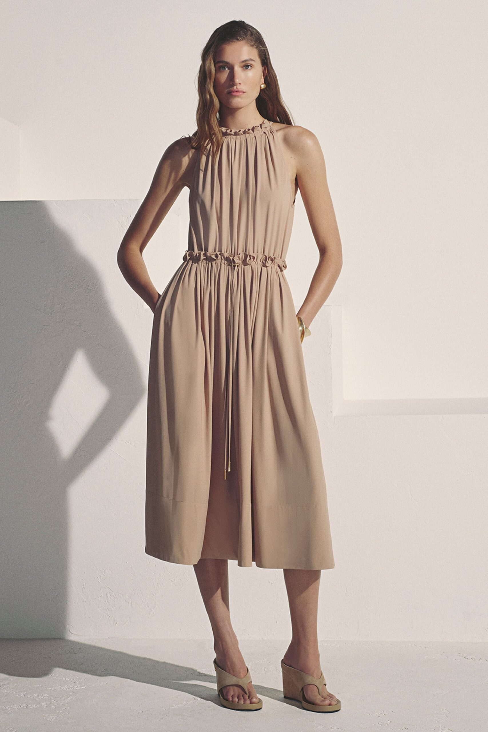 Sand halter midi dress in fluid crepe, ruched at neck and waist. Single round gold earring. Translucent resin bangle at one wrist. Tone-on-tone thong wedges with blunt square soles. Afternoon heat trapped in a whitewashed villa courtyard.