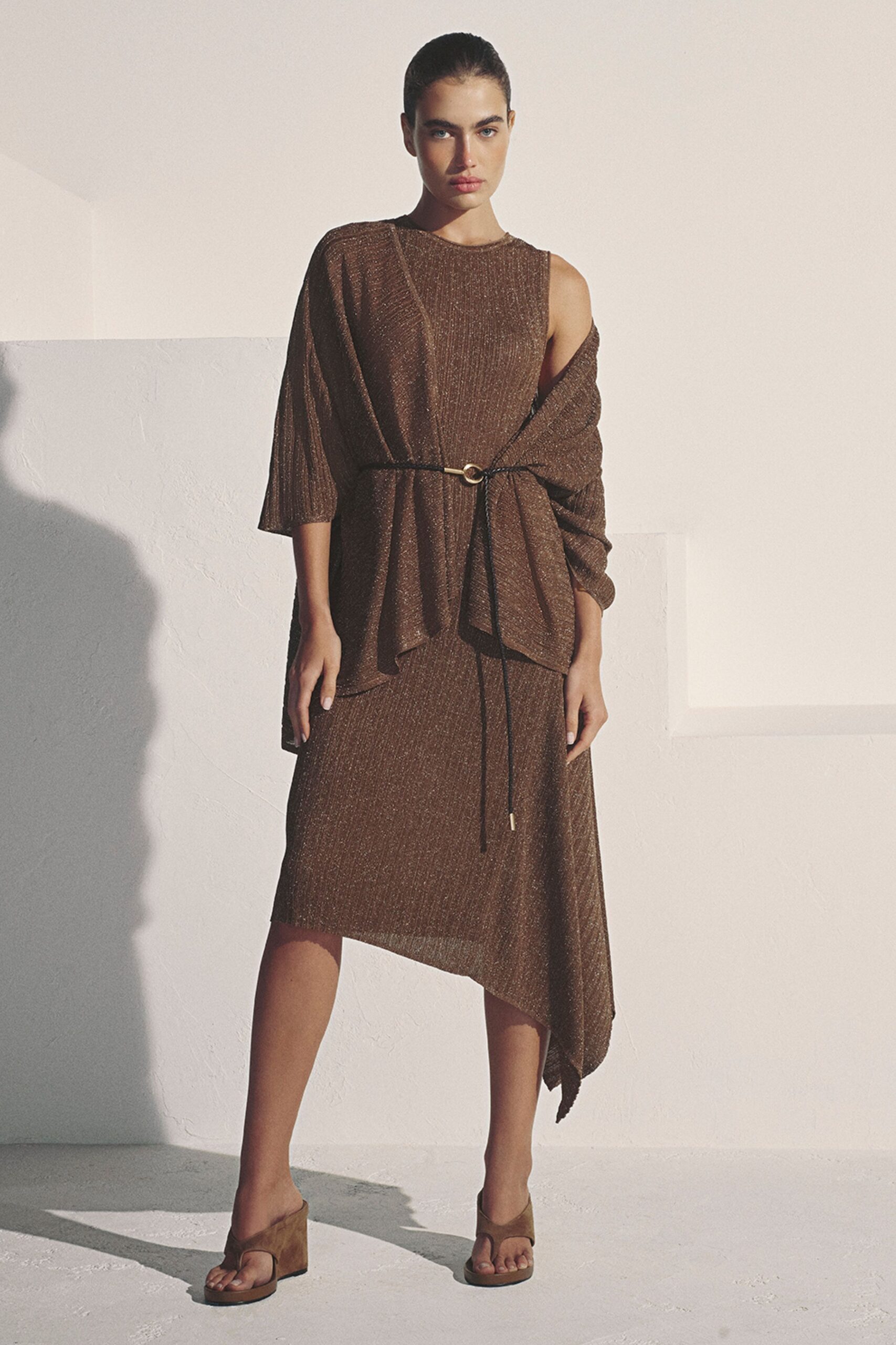 Bronze ribbed knit top with one cape sleeve slipping off the shoulder. Black cord belt caught through a single gold ring. Matching metallic knit skirt, falling to a sharp handkerchief point. Brown suede wedge thong sandals. For terrace drinks before the heat leaves the stone walls.