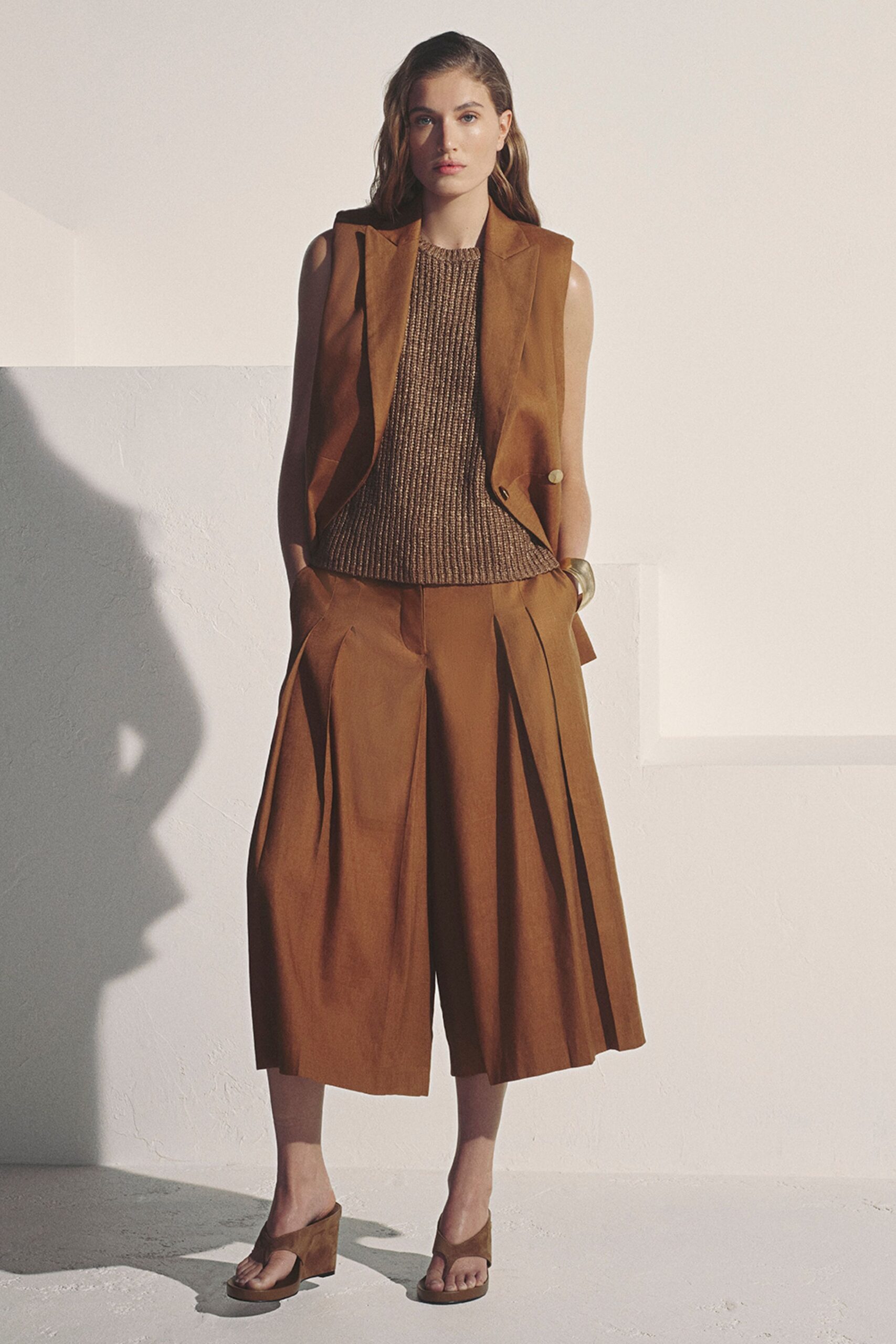 Tobacco suiting vest cut open at the armhole, one horn button low. Brown chunky knit shell with a dense, pebbled rib. Matching wide culottes with deep front pleats breaking into a swing. Brushed gold cuff. Suede thong mules on a block heel. Late-afternoon heat bouncing off stucco after a long lunch ran over.