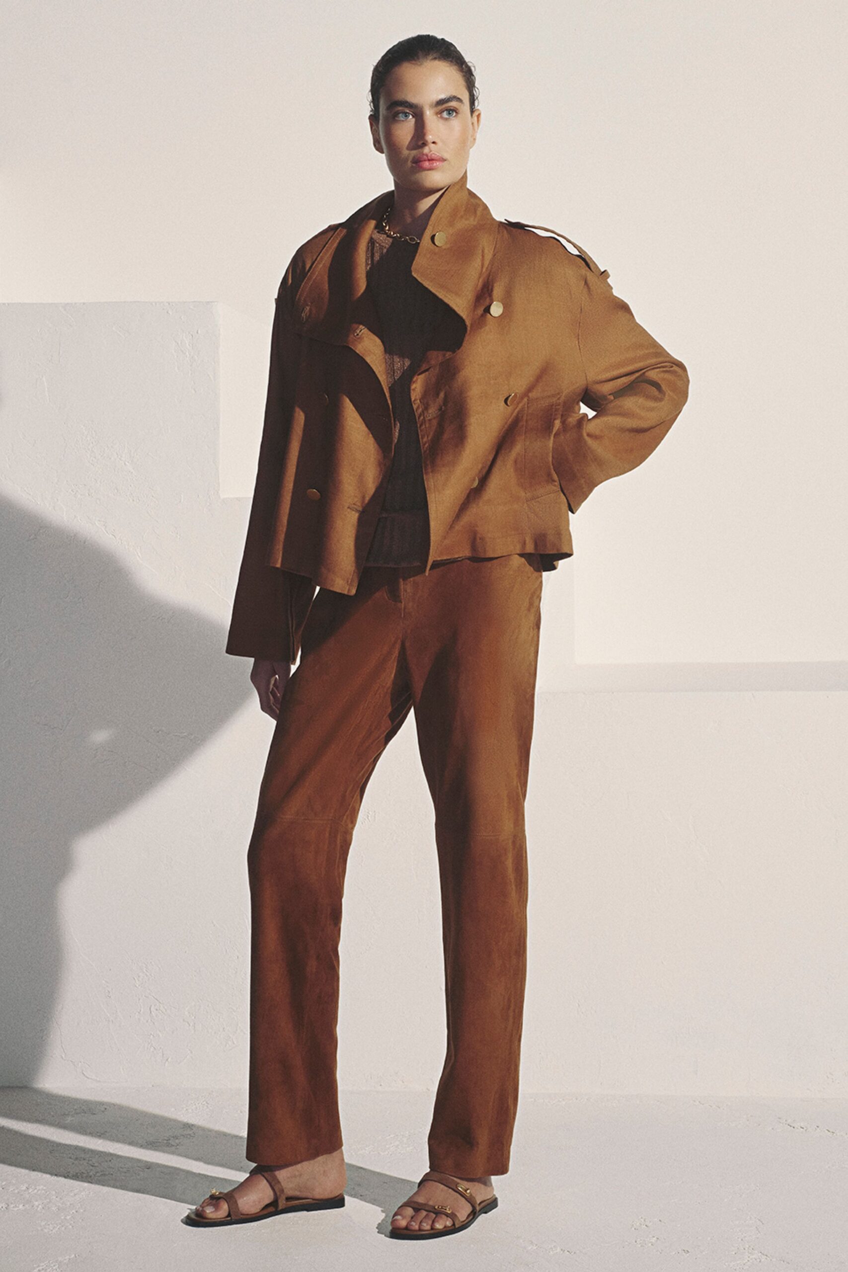 Cropped tan suede jacket cut like a trench, oversized collar kicked open. Black sheer lace top, scalloped at the throat under a chain necklace. Matching suede trousers, long straight leg with a softened crease. Flat brown sandals with small gold rings across the toes. For crossing a sunstruck courtyard before the meeting starts.