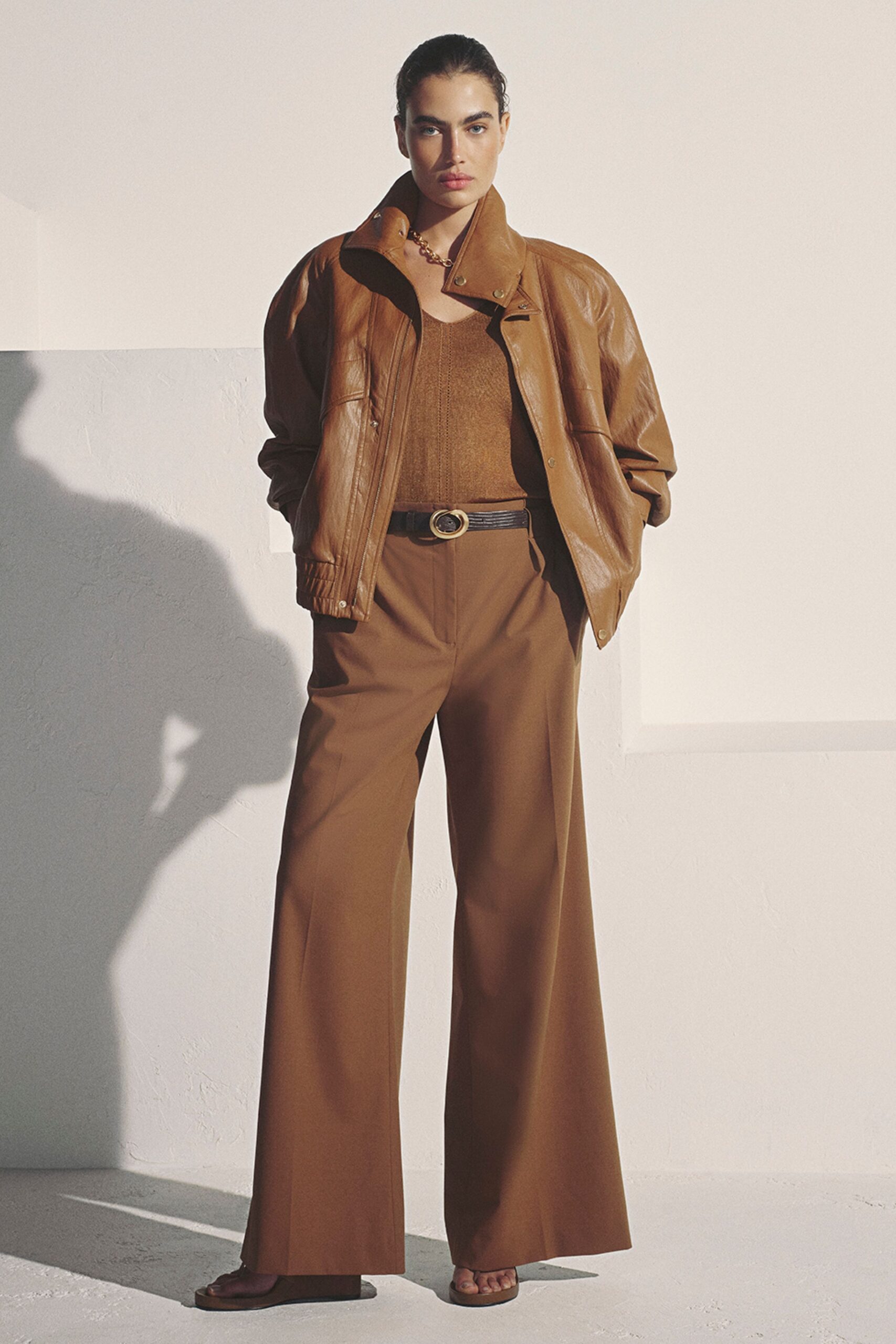 Cognac leather bomber with a high snapped collar and bloused sleeves. Fine brown knit tank with a narrow center seam. Black ridged belt with an oval brass buckle. Tobacco wide-leg trousers pressed with a sharp front crease. Flat brown leather sandals, toes exposed. Sun hitting the hotel corridor before anyone else is downstairs.