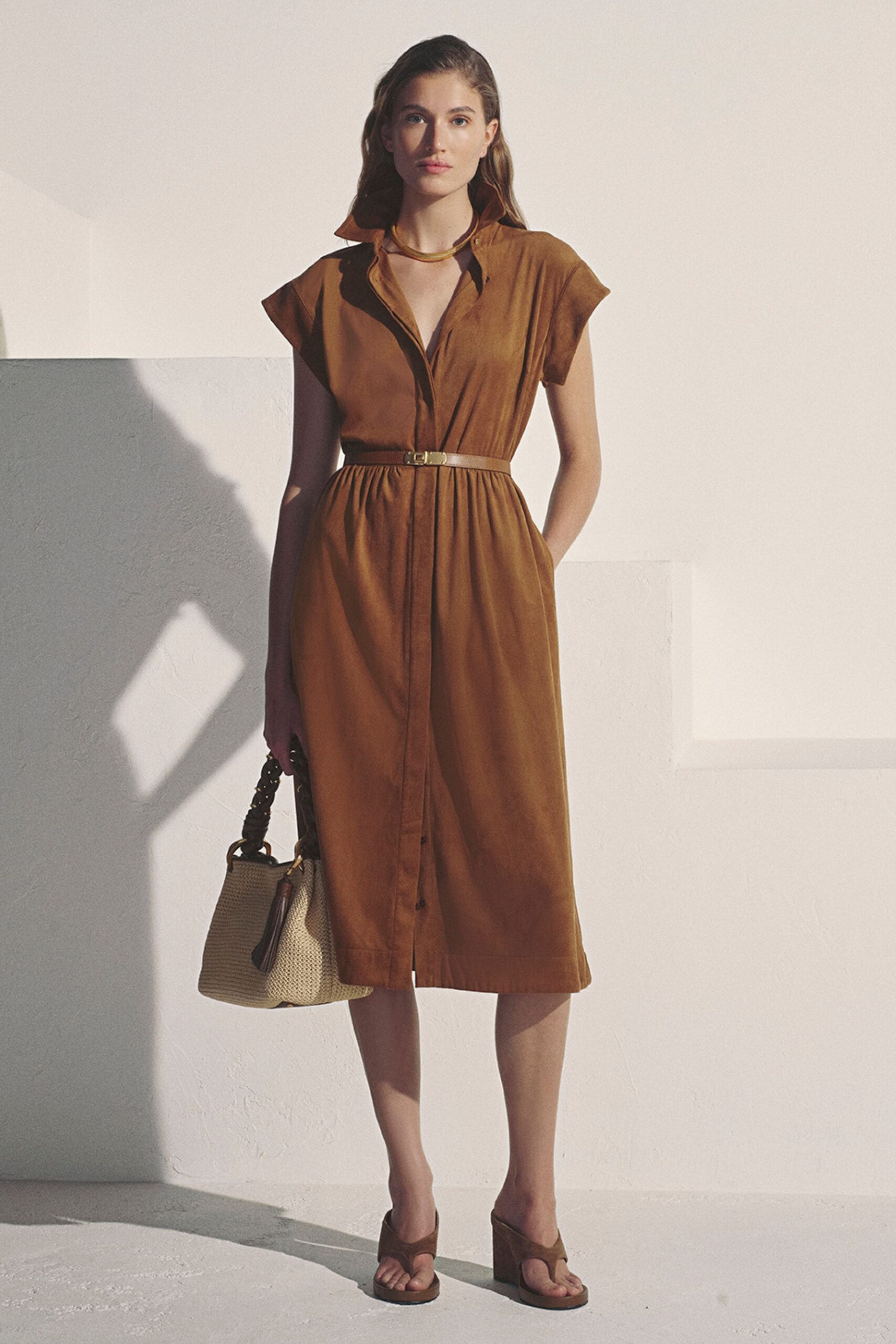 Curved gold collar sitting close to the base of the throat. Cinnamon shirt dress in suede-like cloth, softly gathered under a skinny belt. Cream woven tote with a twisted handle and long tassel. Brown thong sandals with thick platform soles. For a hotel terrace lunch gone quiet in the heat.