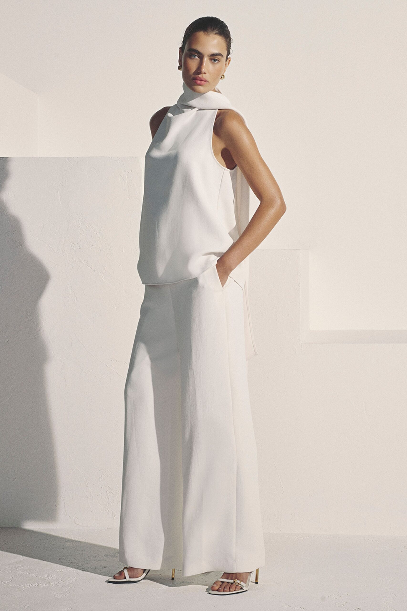 Ivory crepe shell with a scarf collar folded close at the throat. Matching full-leg crepe trousers with a clean front crease and side ties. White thong sandals on slim gold stiletto heels. Small gold drop earrings. Sun pooling across a plaster villa before the lunch guests arrive.