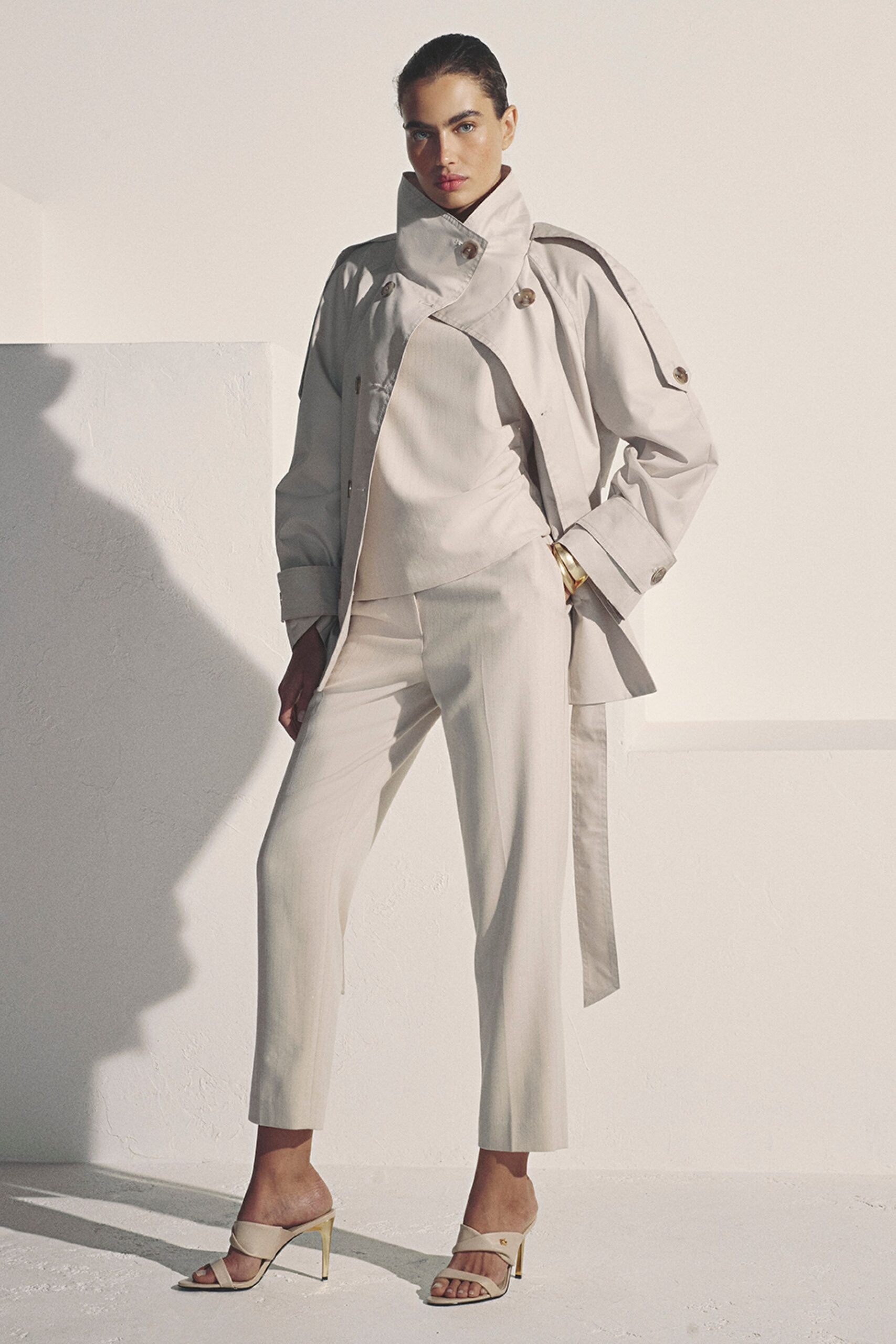 Chalk cotton trench with a tall funnel collar and sleeve-tab buttons. Ivory wrap top, ruched tight through one side of the waist. Slim cropped trousers in the same dry suiting cloth. Cream cross-strap mules on thin gold-toned heels. First appointment after moving somewhere with harsher light.