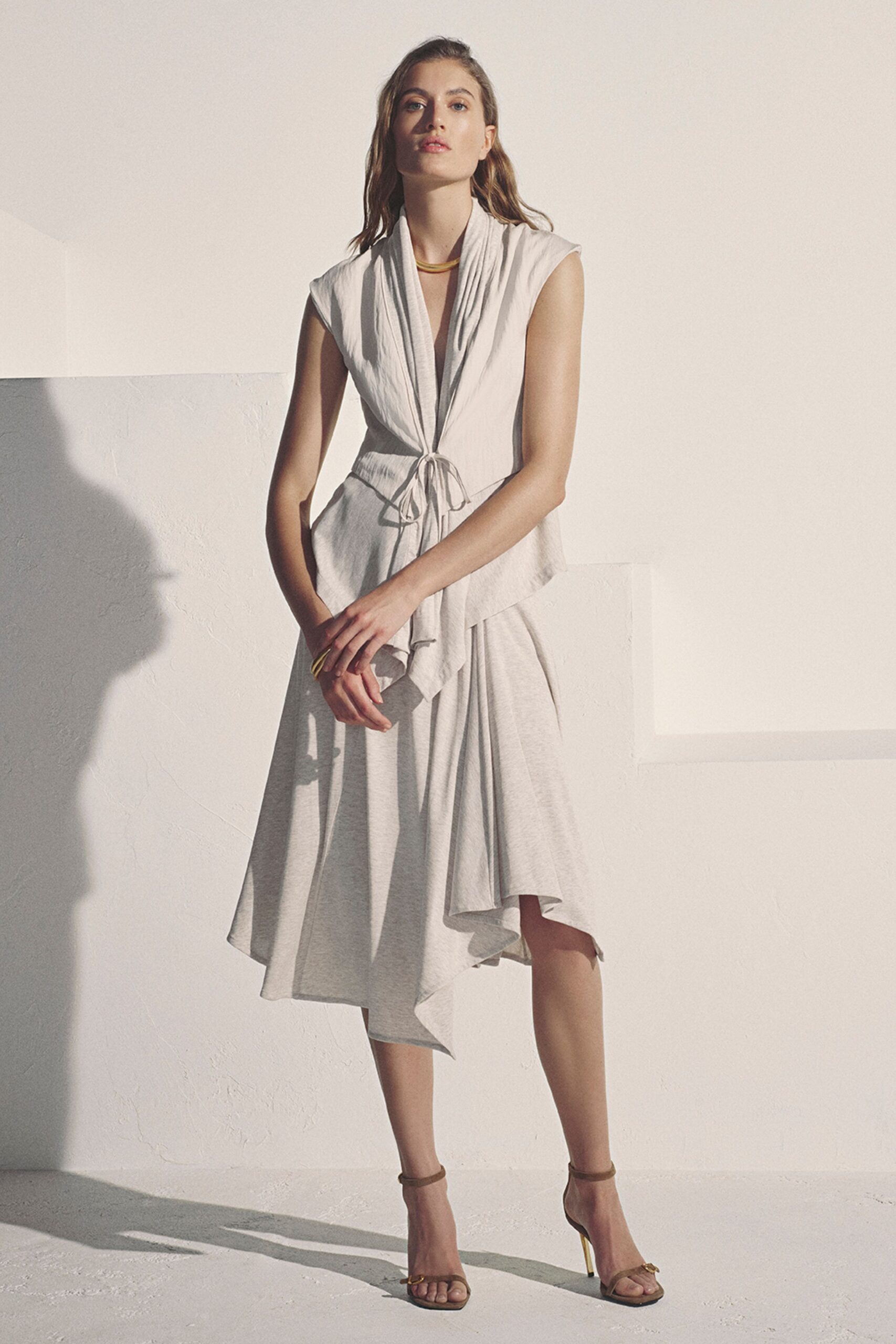Pale heather jersey top with a draped shawl collar tied at the waist. Matching asymmetrical skirt, one point dropping lower at the right shin. Curved gold collar close to the throat. Taupe ankle-strap sandals with slim gold stiletto heels. Late-afternoon terrace party, white walls still holding the day's heat.