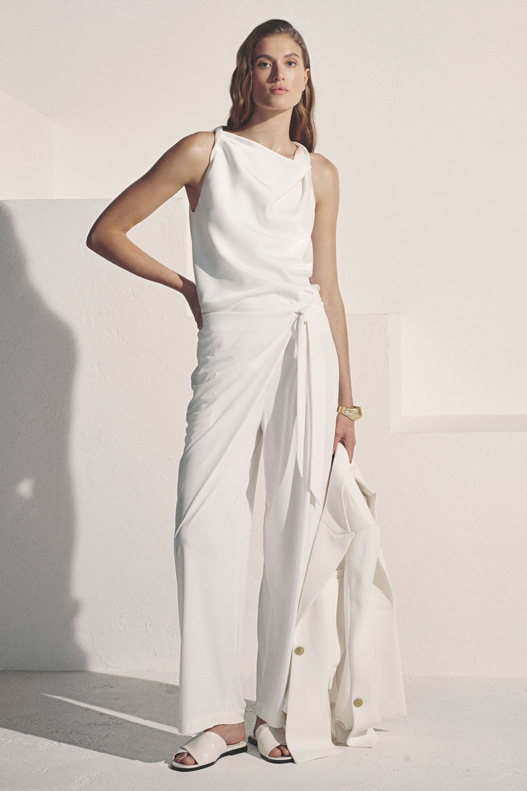 Ivory crepe top with a soft draped cowl crossing one shoulder. Matching wrap-front trousers, tied at the waist with side ruching. Brushed gold cuff. White padded slide sandals. Long ivory coat carried in hand, oversized gold snaps at the hem. For the hotel terrace just after the wedding crowd has gone upstairs.