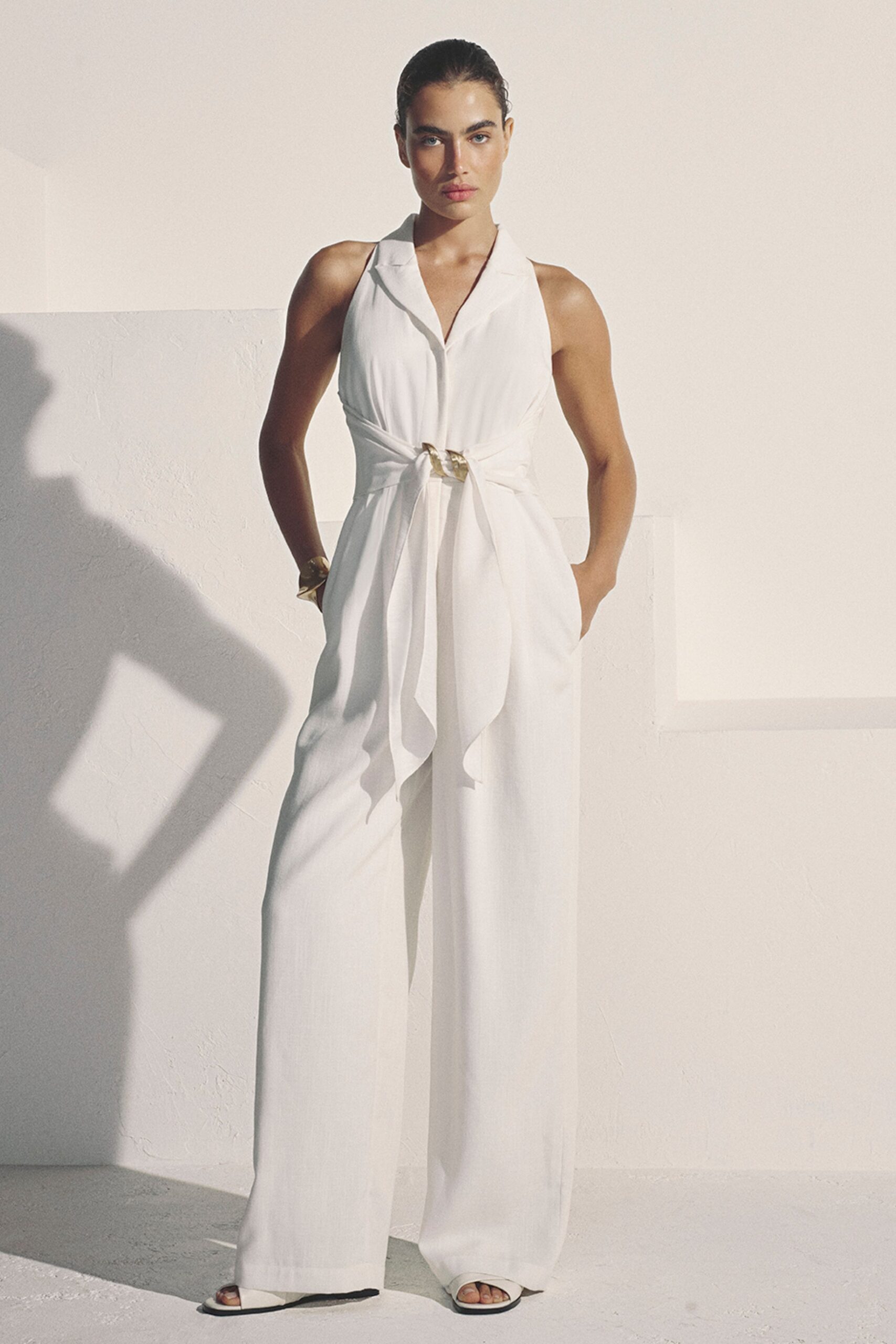 Ivory halter jumpsuit with a notched collar and sash threaded through a gold ring. Crinkled gold cuff at the wrist. White slide sandals with squared toes peeking past the hem. Sun hitting the hotel arcade before anyone has claimed the loungers.