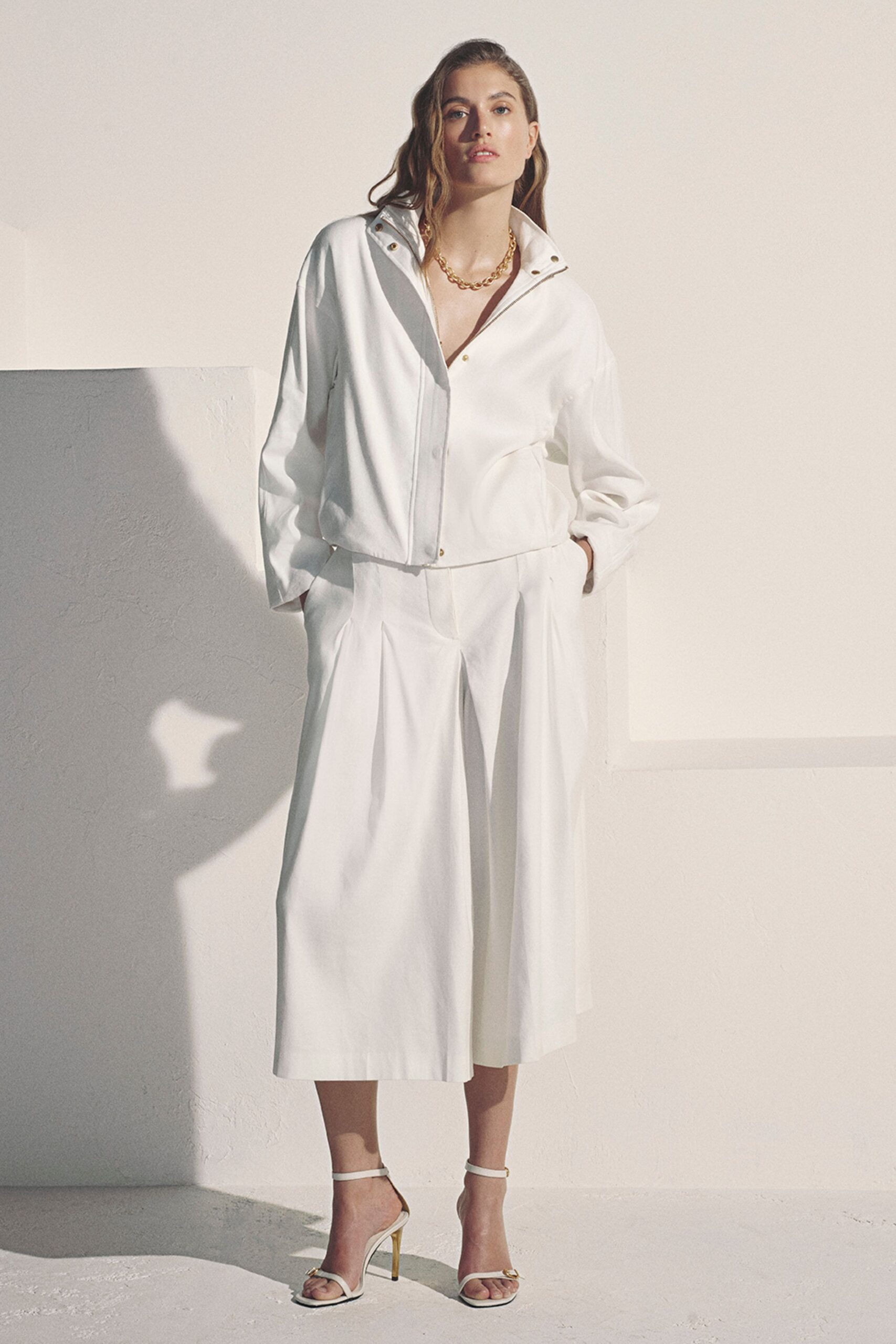 Heavy gold curb chain at the open throat. Chalk drill jacket with brass snaps climbing the stand collar. Matching culottes cut wide with deep front pleats. White ankle-strap heels with a small gold toe bar. Morning light in a rental with the windows already open.