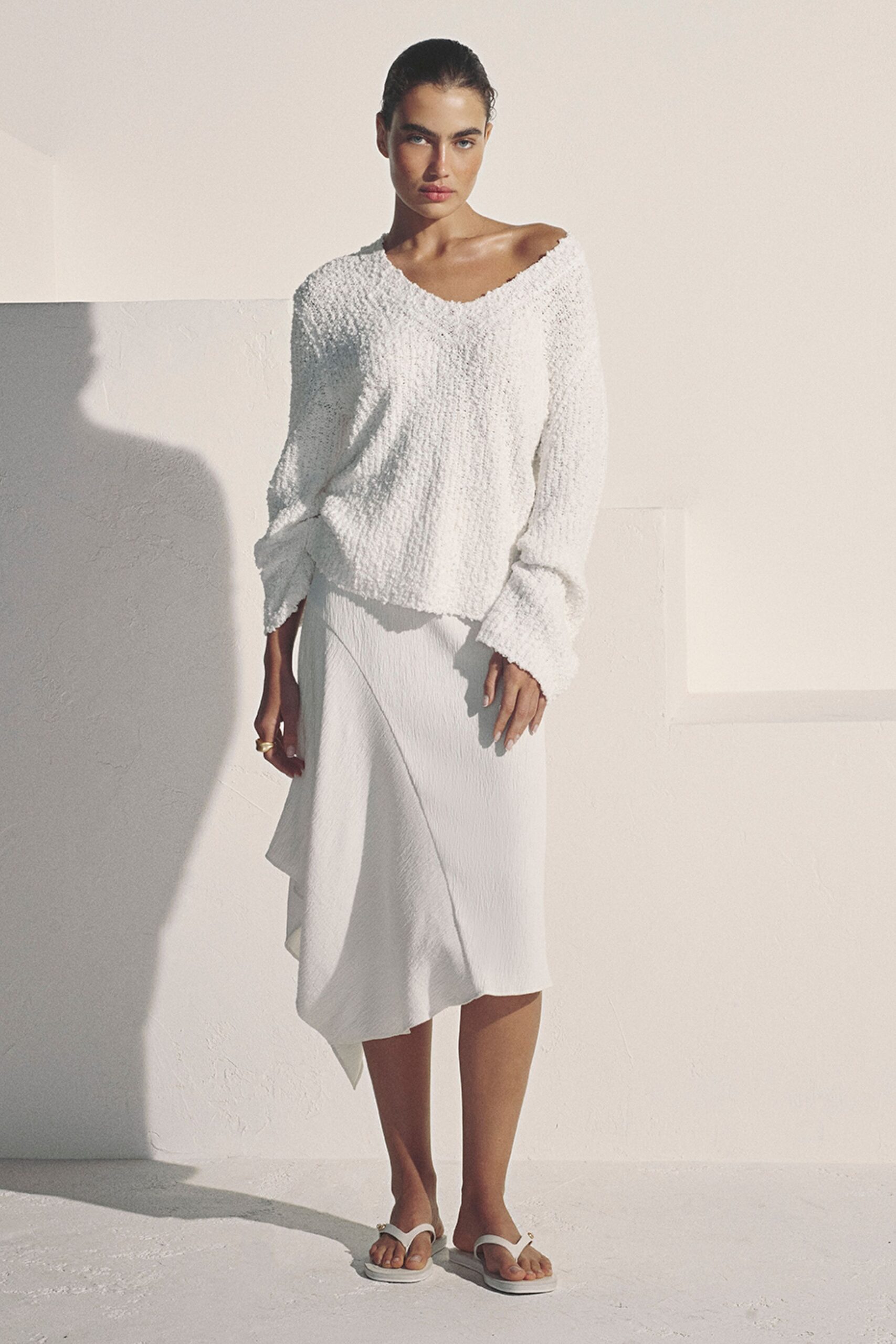 White boucle sweater with a wide torn-edge neckline slipping off one shoulder. White crepe midi skirt, cut on the bias with an angled handkerchief hem. Chunky gold ring. White thong sandals with a tiny gold stud at the strap. Sunlight on limewashed walls, the hotel room still cool after an early swim.