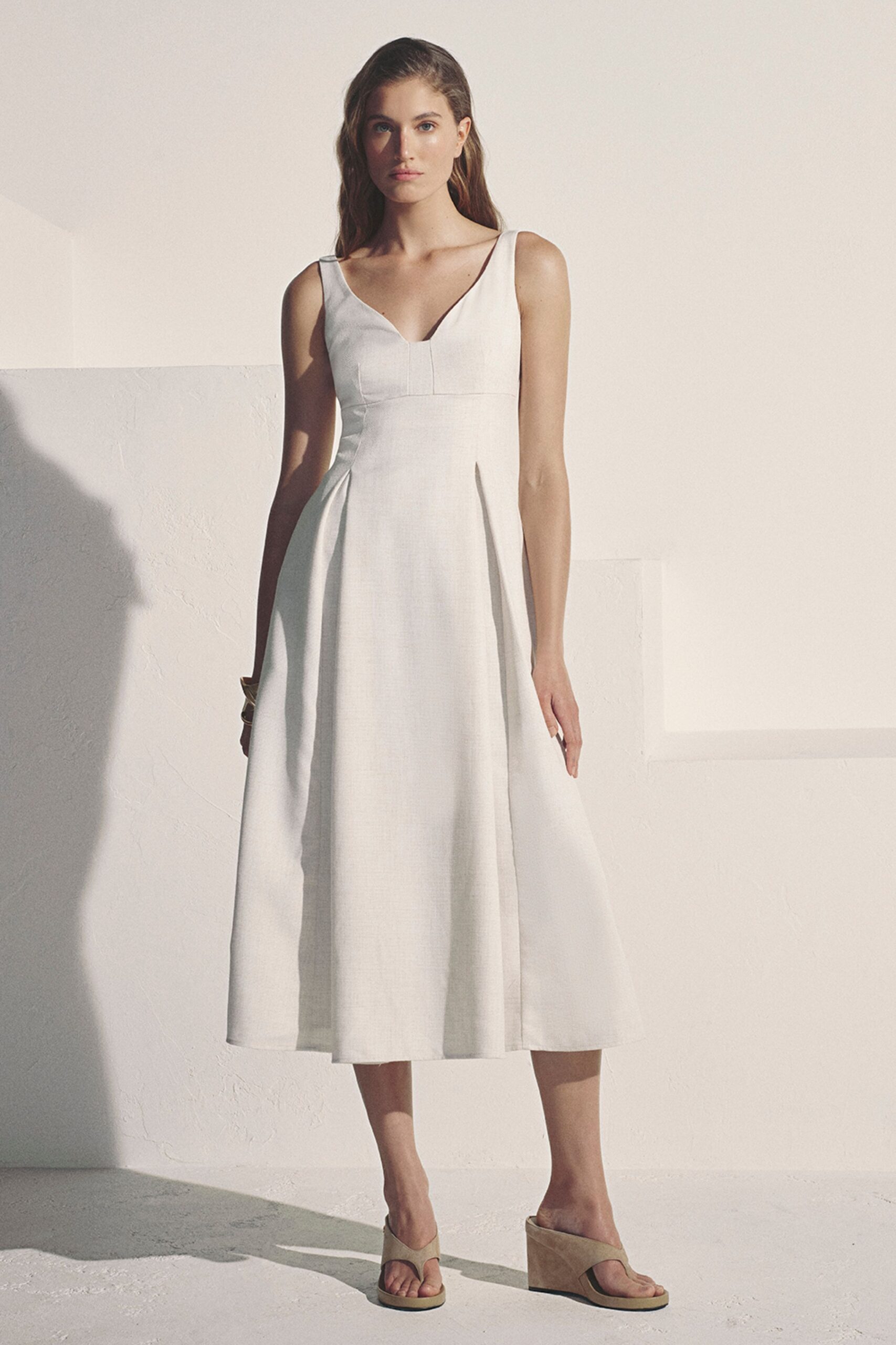 Ivory sleeveless midi dress in crisp suiting cloth, paneled V bodice and deep skirt pleats. Bone suede thong wedges with a thick platform sole. Terrace wedding at four, heat still rising off the stone.
