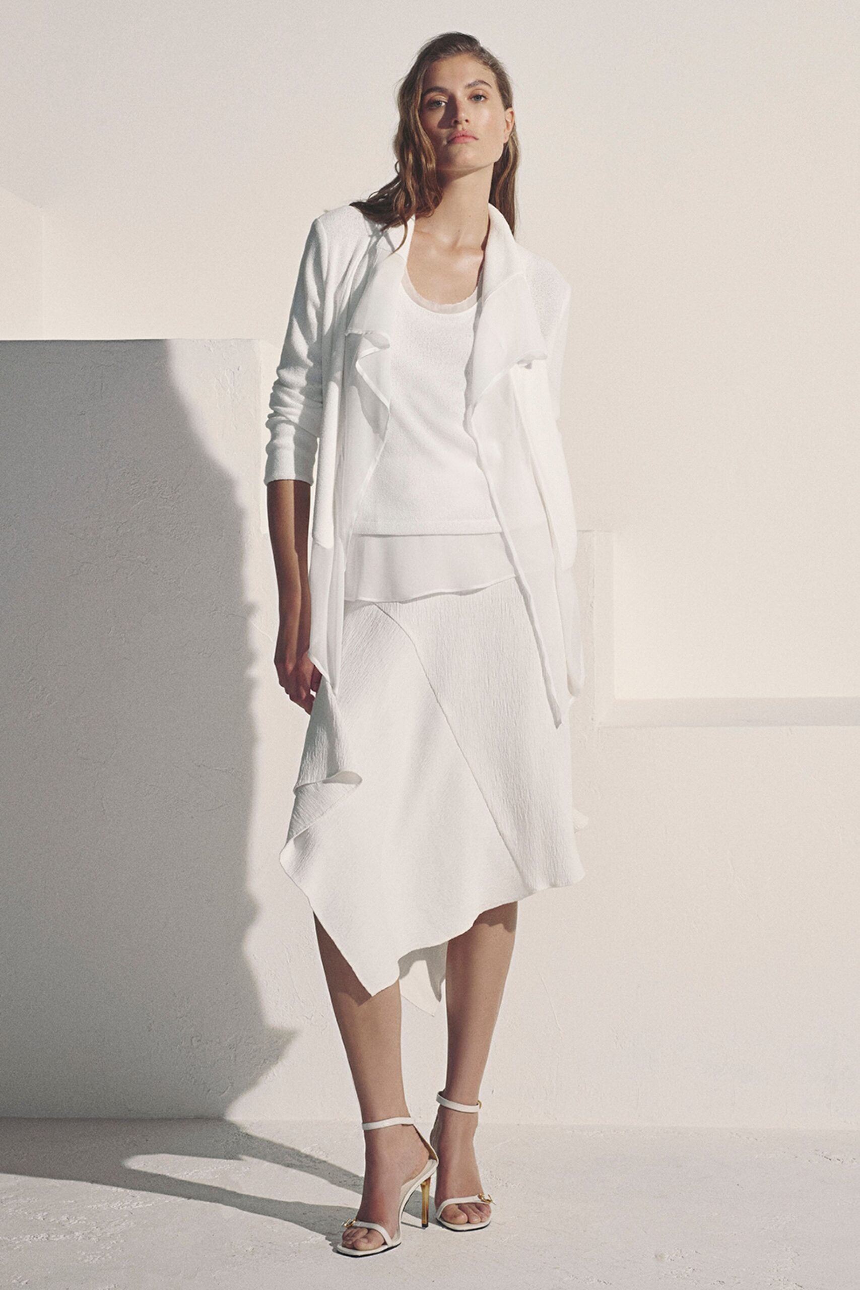 Open white jacket with softly folded lapels and a long uneven front. Fine white knit shell, the scoop neck lightly rolled. Crinkled white wrap skirt cut with a floating handkerchief hem. White ankle-strap sandals with slim metallic heels. Sun on a hotel terrace before everyone else has come downstairs.