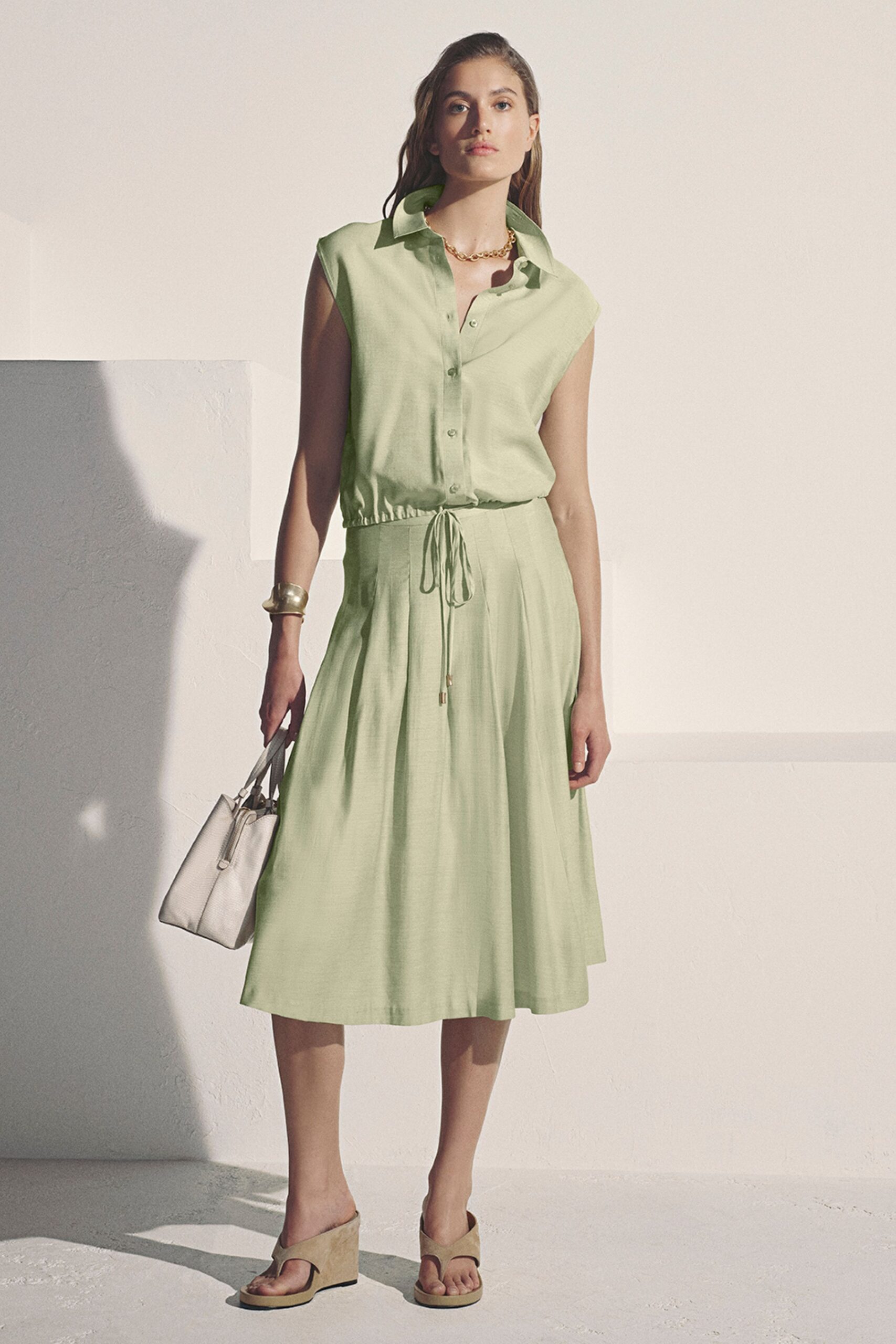 Pale pistachio shirt dress with cap sleeves and a drawcord waist, metal tips hanging low. Short gold chain links at the open collar. Wide brushed-metal cuff. White pebbled-leather tote with slim handles. Taupe suede thong wedges. Hotel courtyard at noon, carrying the room key and yesterday's paperback.