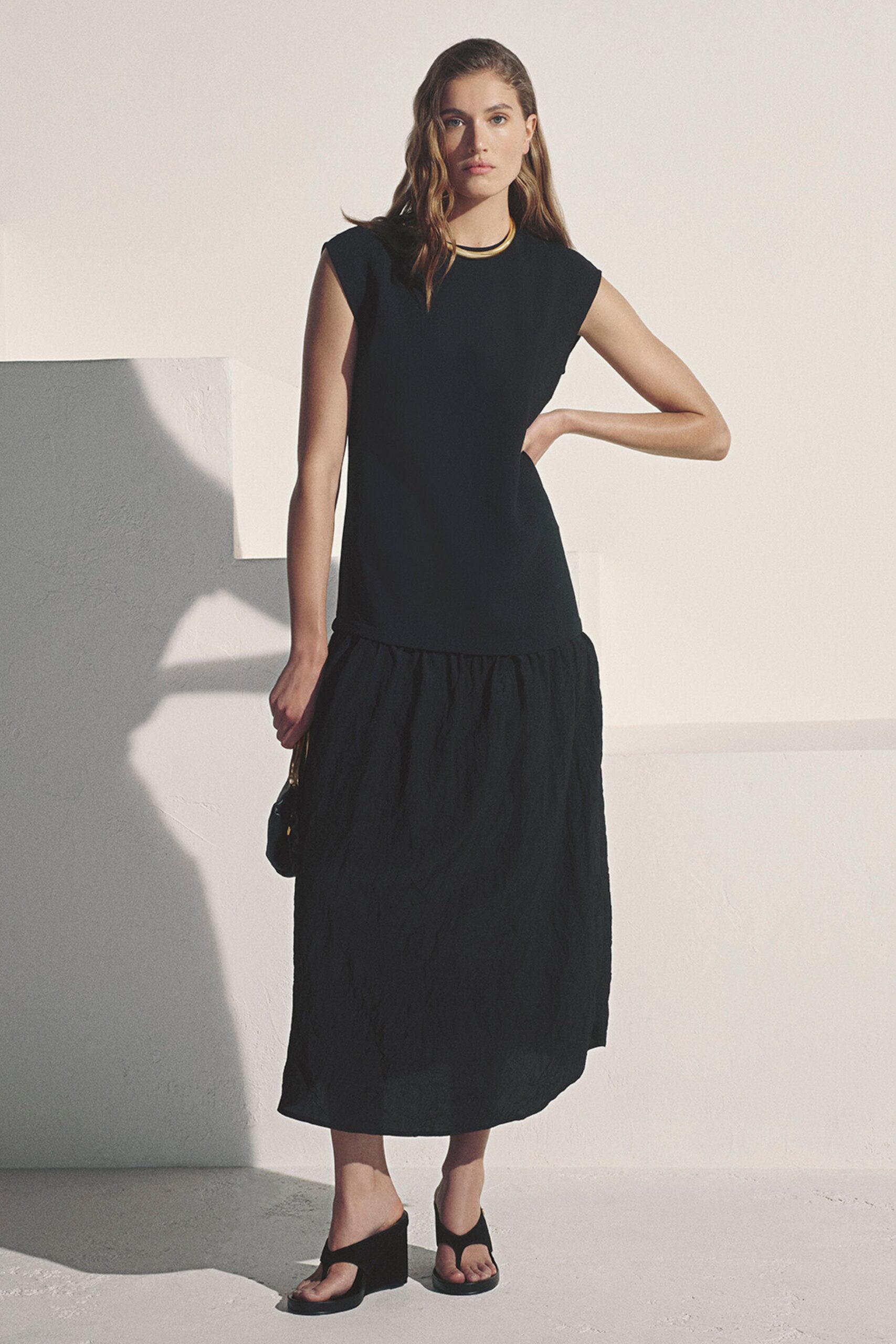Rigid gold collar sitting close at the base of the throat. Black sleeveless column dress with a crinkled drop-waist skirt panel. Small black shoulder bag with a glinting gold strap ring. Black thong wedges with thick soles. Walking across a whitewashed terrace just before the heat lifts.