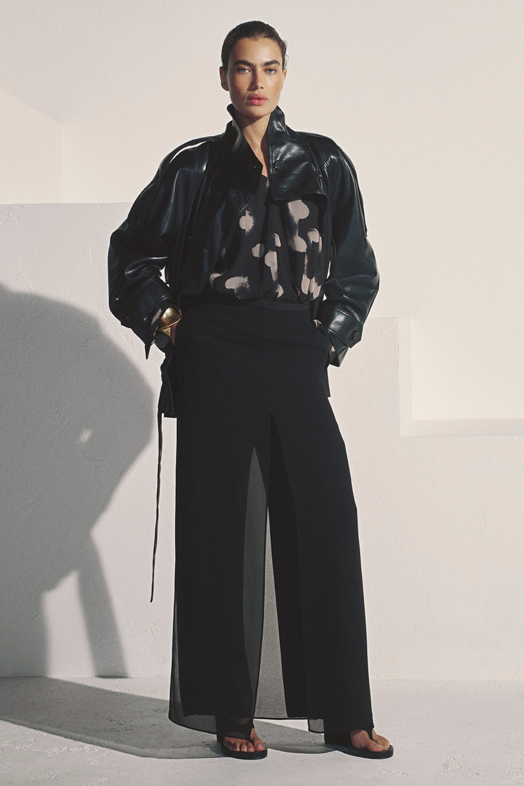 Black coated jacket with a high stand collar and loose hanging ties. Inky draped top, blotched with pale cloudlike spots. Black wide-leg trousers split to show a sheer inner panel. Brass cuff at one wrist. Flat black thong sandals. Walking into a whitewashed villa after sunset, glass sweating in your hand.