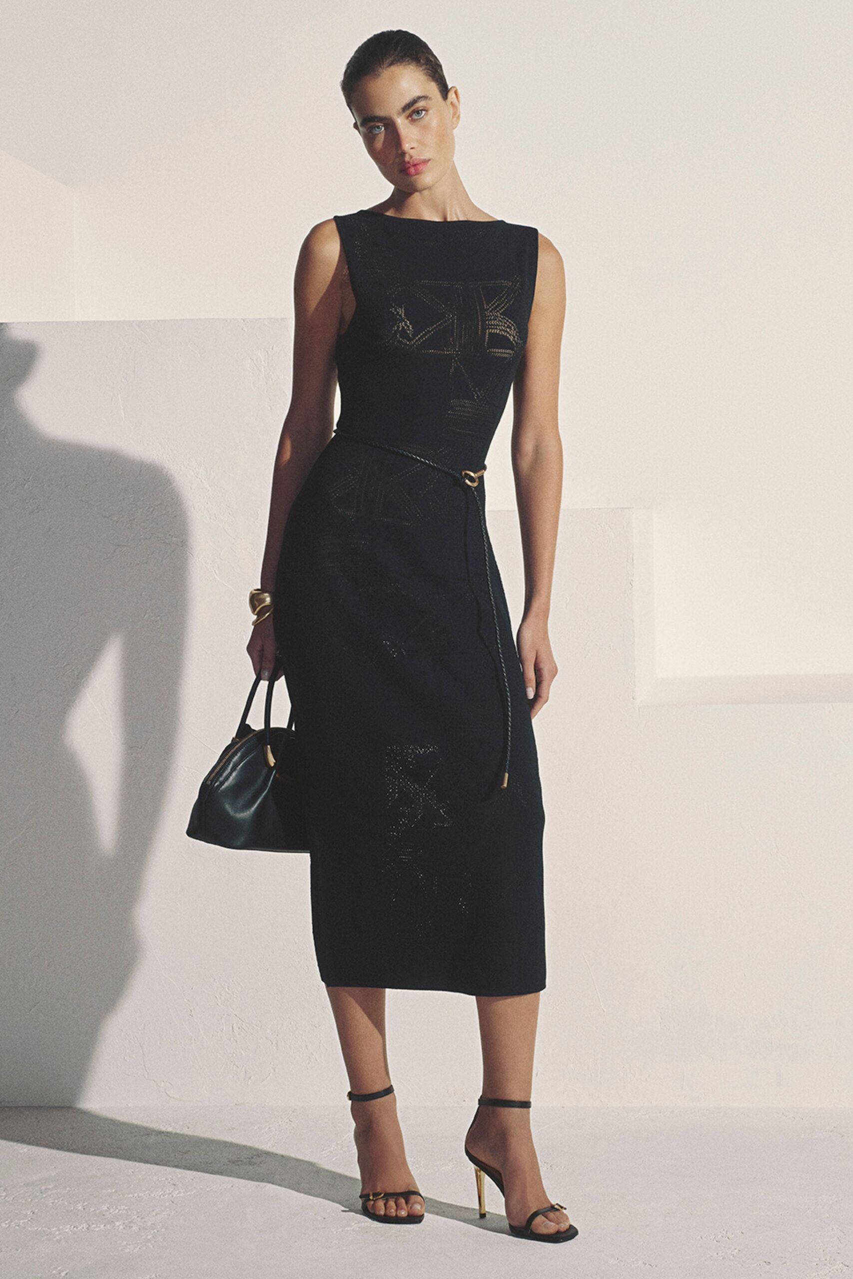 Black sleeveless knit midi dress with sheer geometric panels and a rope belt. Chunky gold cuff, folded like soft sheet metal. Black leather top-handle bag with a crescent brass side piece. Barely there black sandals on thin gold stiletto heels. For terrace drinks just before the light drops off the stucco walls.