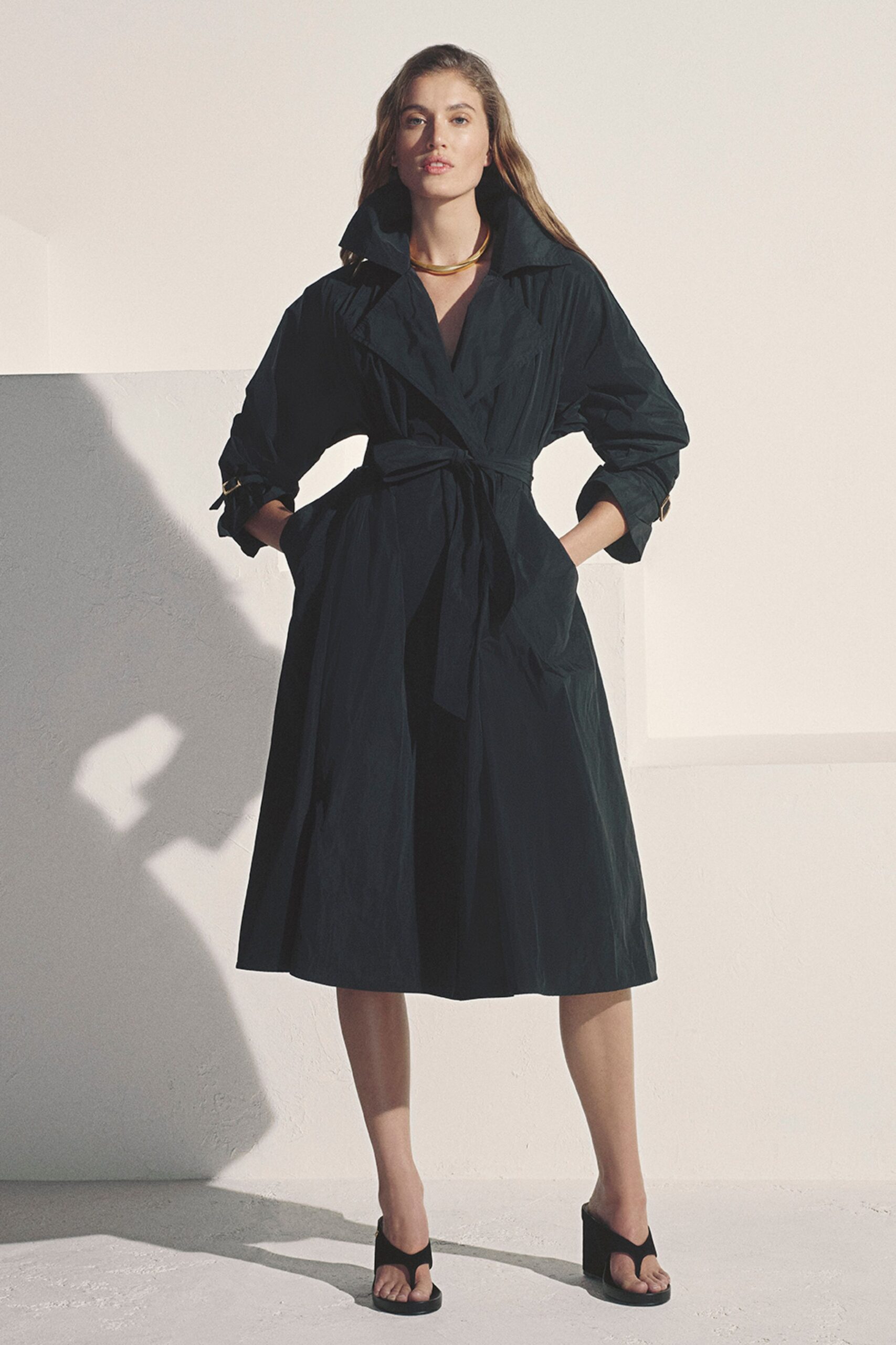 Rigid gold collar sitting close above the open throat. Ink-dark cotton coatdress with a popped collar and self-tie belt. Cuffs cinched by tiny strap tabs at the forearm. Black thong mules on blunt block heels. Crossing a whitewashed courtyard before dinner while the stone still gives back heat.