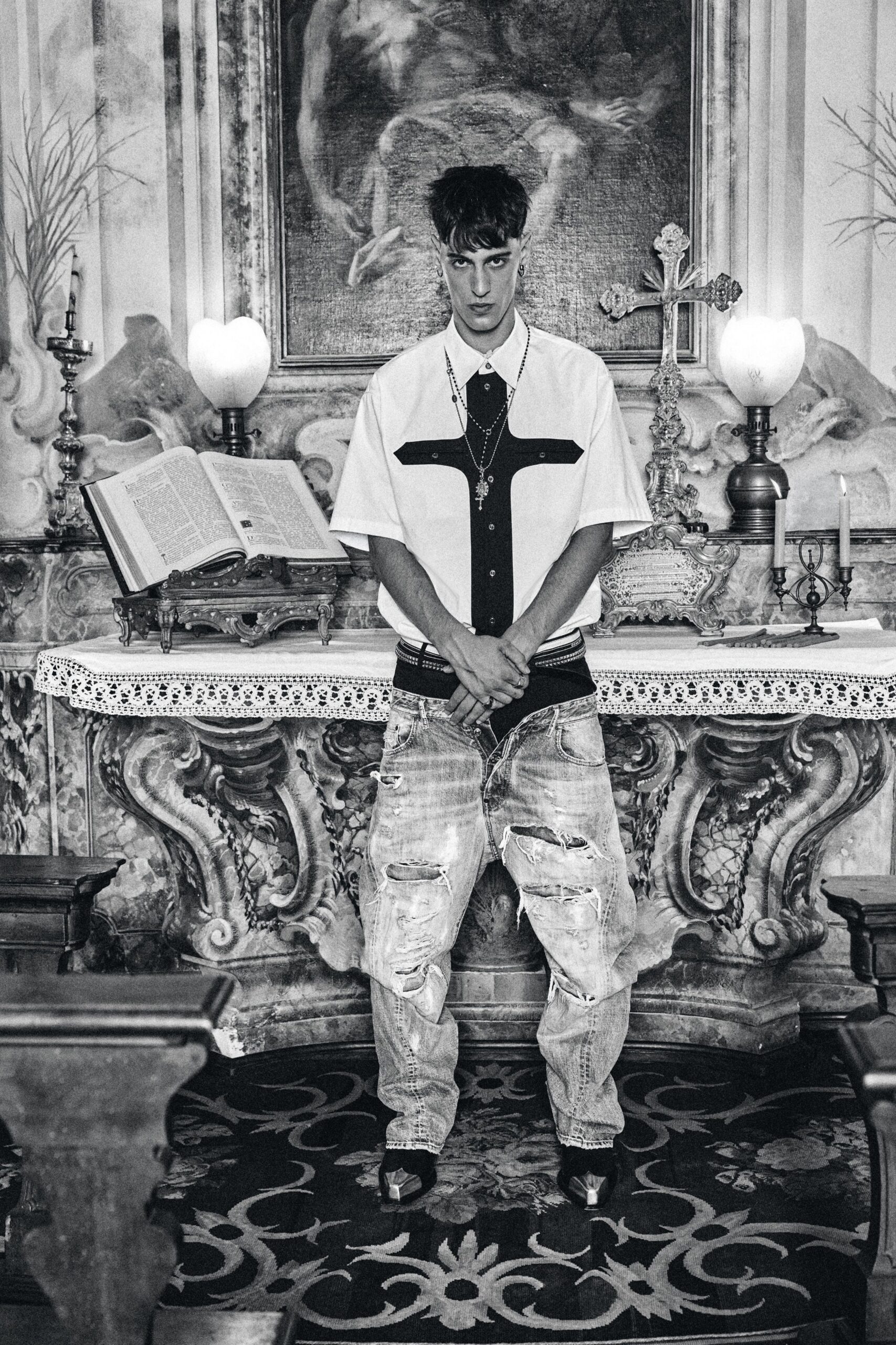 White short-sleeve shirt with a black cross-shaped placket yoke. Layered rosary chains dropping over the chest. Studded double belt peeking above the waistband. Bleached jeans shredded open at both thighs and knees. Pointed boots with metal toe caps. For lighting candles in a side chapel after missing the last train.
