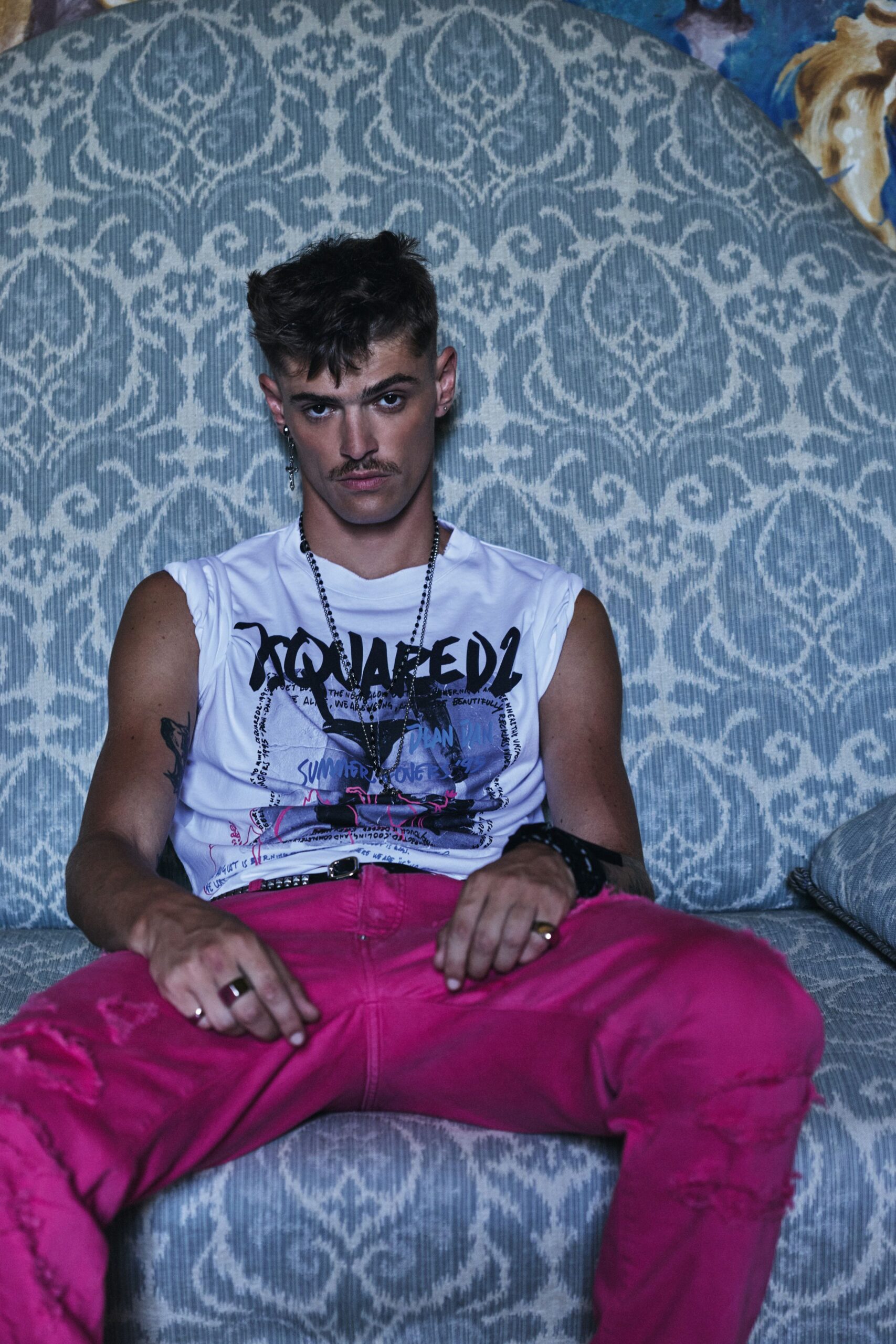 White jersey sleeveless tee with a dense black-and-pink graphic print. Layered gunmetal chains falling over the chest. Narrow black belt dotted with silver studs. Hot-pink jeans with ripped knees and a raw frayed tear. Sinking into a hotel sofa after the gig, still wired and not going upstairs yet.